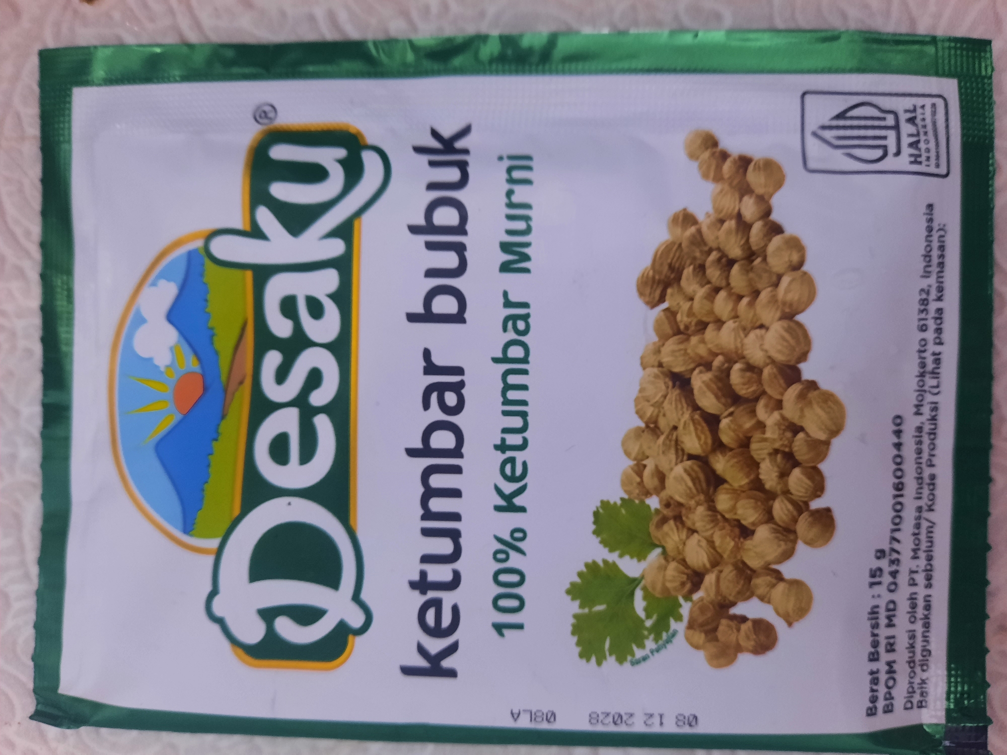 The image displays the front of a packet of "Desaku Ketumbar Bubuk" (Desaku Coriander Powder). The packaging features a green border with white and green accents. Prominently displayed is the brand logo "Desaku" in large, stylized white letters with a yellow outline, set against a backdrop of a stylized landscape with mountains and a sun. Below the brand name, the product name "ketumbar bubuk" is written in dark blue, followed by "100% Ketumbar Murni" in green. A photograph of coriander seeds and a sprig of coriander leaves is situated at the bottom of the packaging. Text indicating the net weight, BPOM registration number, production details, and a "Halal Indonesia" logo are visible at the lower section of the packet. The packet is placed on a textured white surface. There is no indication of the city of Lhokseumawe, Indonesia, in the image.