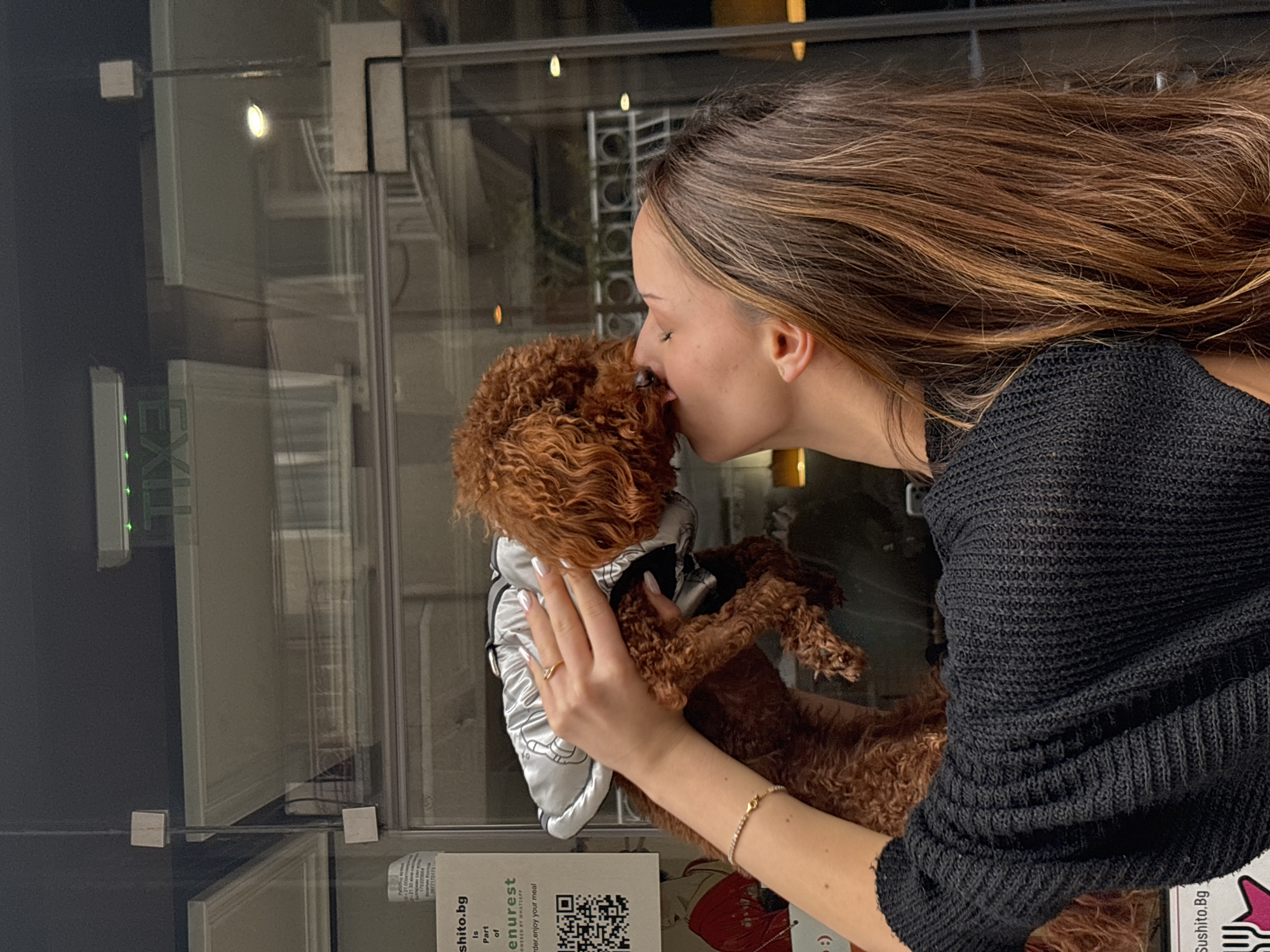 A woman with long brown hair is holding a brown poodle wearing a silver jacket and kissing the poodle's nose. The woman is wearing a black sweater and a delicate bracelet on her left wrist. She also has a ring on her finger. They are standing next to a glass wall that has an "EXIT" sign visible, along with some restaurant advertisements for "sushito.bg" and "menurest" which identifies the location as likely being in Sofia, Bulgaria.