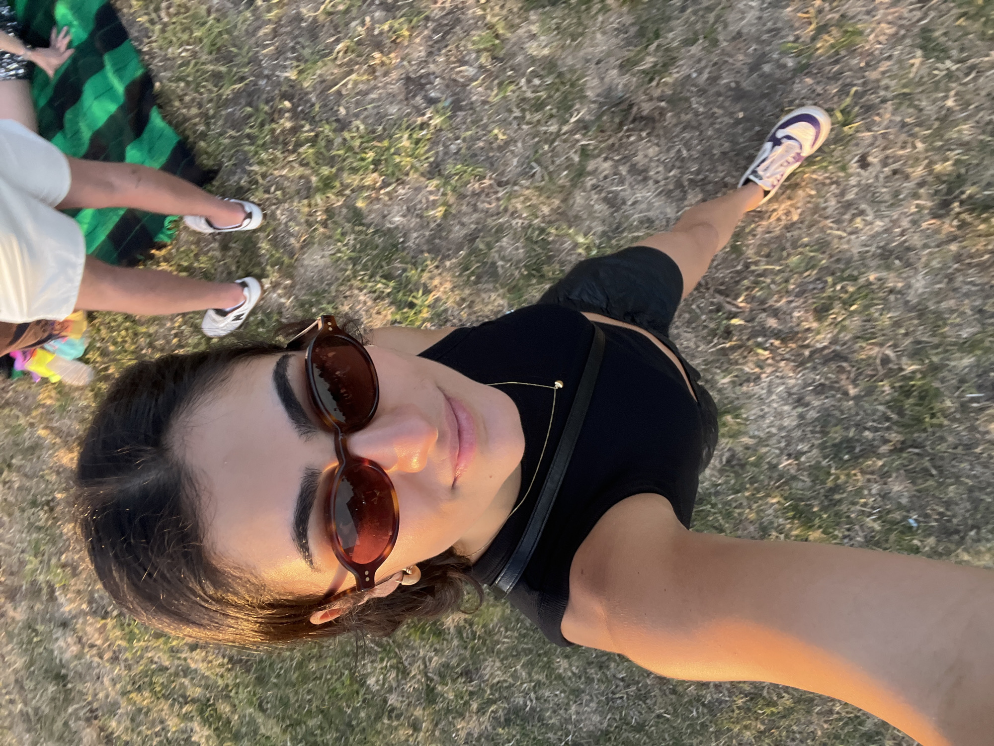 The image shows a woman from a high-angle perspective, taking a selfie outdoors. She is wearing a black crop top, black shorts, and white sneakers with purple accents. She has on tortoiseshell sunglasses and a gold necklace. She is standing on dry grass with some green patches. In the background, to the right, are the legs of two other people, one wearing white shorts and the other black and white patterned shorts, sitting on a green and black striped blanket. The lighting suggests it is late afternoon or early evening, possibly during golden hour. The environment appears to be a park or an open grassy area in Porto, Portugal.