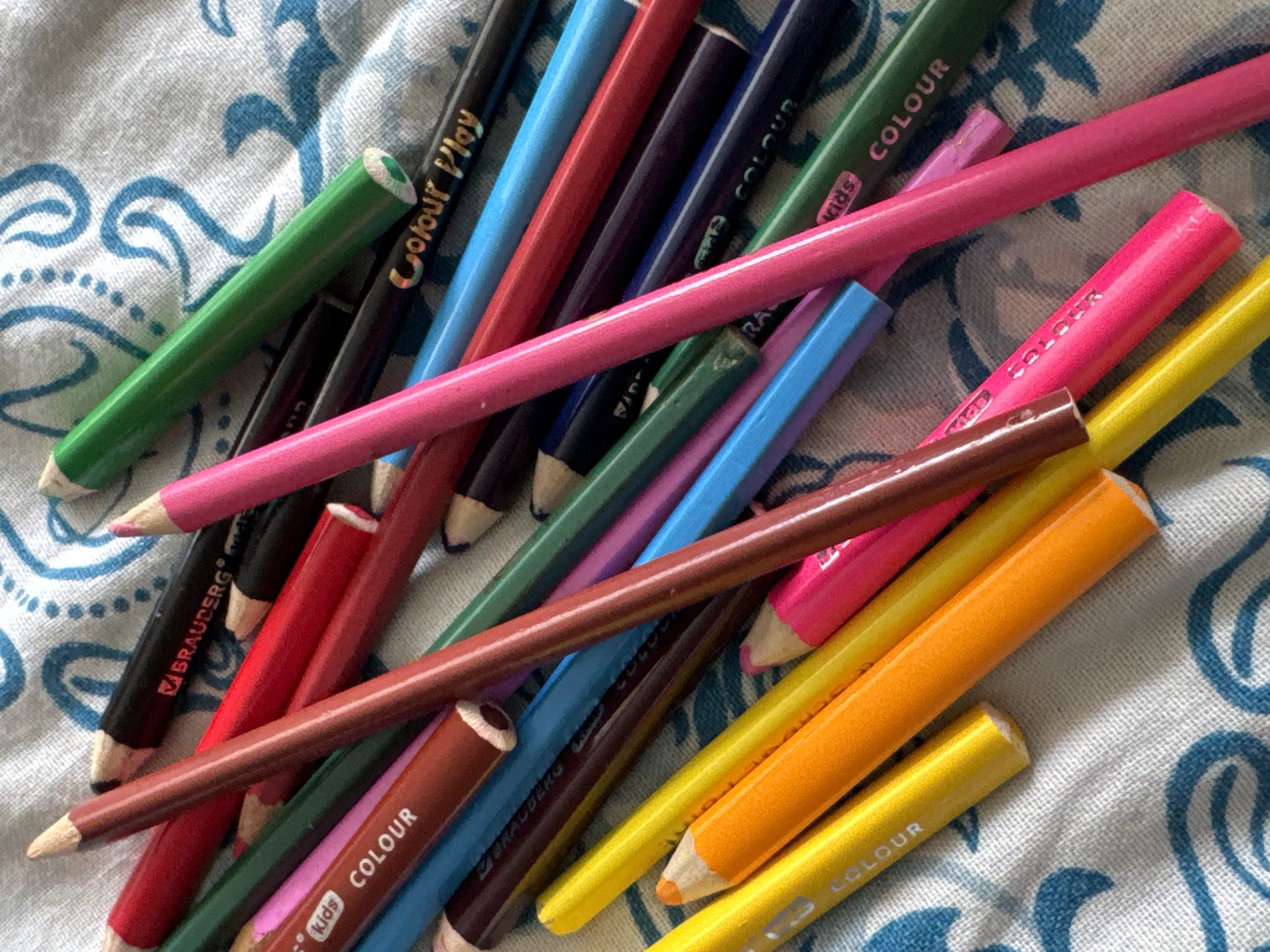 A close-up, overhead shot shows a pile of colored pencils on a textured white and blue patterned fabric. The pencils are of various colors, including red, black, green, pink, orange, yellow, brown, and blue, and they are arranged in a disorganized manner, overlapping each other. Some pencils have visible branding, such as "BRAUBERG" and "Colour Play." The fabric appears to be a tablecloth or bedspread with a repeating wave-like blue design. The image does not depict any specific location within Яромінскі сельскі Савет, Belarus, or any people or actions.
