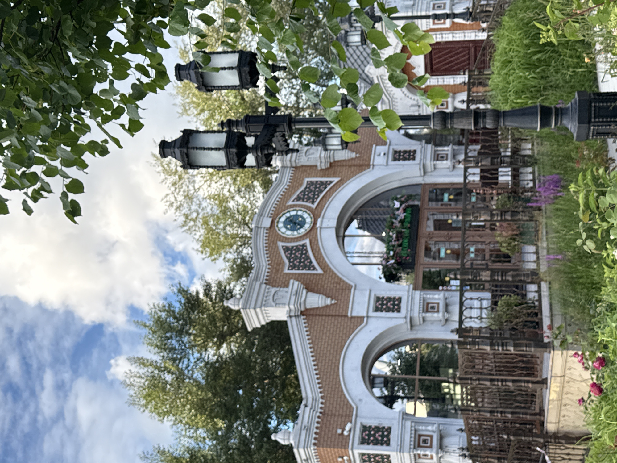 This is a vertically oriented, eye-level medium shot of an ornate, arched entranceway, possibly a building facade or a gate, viewed from an angle. Lush green foliage from trees overhangs the scene from the upper left, with a cloudy blue sky peeking through. Two large, black, lantern-style light fixtures are mounted on poles above the entrance.
The entrance itself is detailed with a combination of white plasterwork and reddish-brown brick. It features a prominent circular clock with Roman numerals set within a decorative surround. The archway opens into a space with glass windows and what appears to be decorative fencing or railings.
Behind the entrance, glimpses of interior elements are visible, including shelving, greenery, and possibly shops or restaurants. There are also more decorative railings and planters with flowers. The overall impression is one of an inviting and well-maintained public space, likely in a park or a commercial area.
The lighting suggests it could be daytime, with diffused light indicating possibly overcast conditions or the softer light of morning or late afternoon. The location is identified as Moscow, Russia. There is no discernible activity or people visible in the frame. No prominent text is visible, other than what might be small signs within the building's interior, which are too blurry to read.