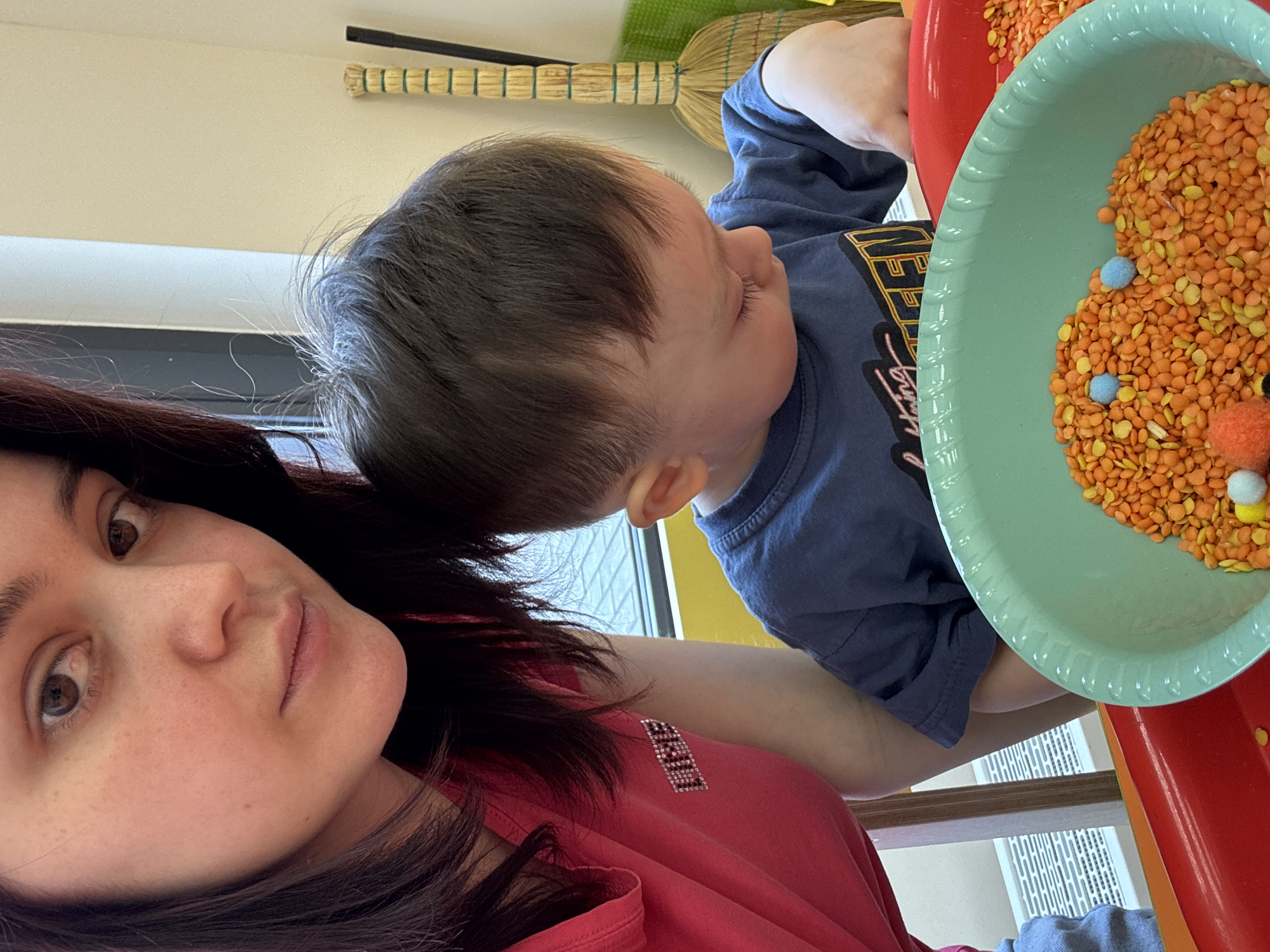 This vertically oriented indoor photograph, taken in Izhevsk, Russia, depicts an adult woman and a young child engaged in a sensory play activity. The image is rotated 90 degrees clockwise, placing the subjects on their sides.
In the foreground, on the left, is the adult woman. She has dark hair and dark eyes, looking with a neutral expression towards the camera. She is wearing a red or deep pink short-sleeved top. A sparkly, possibly rhinestone, patch on her shoulder features some text, which is not clearly decipherable.
To her right, a young child, likely a boy, with dark hair, is visible. He is wearing a blue t-shirt with some yellow graphic design on it, which is also not clearly legible. The child is focused downwards, intently looking at a sensory bin placed on a red surface, likely a table.
The sensory setup consists of at least two shallow plastic dishes: one red and one light blue-green. These dishes are filled with a colorful mixture of orange lentils and lighter yellow/tan dried peas or grains. Several small, fluffy pom-poms, in light blue and orange colors, are scattered among the legumes, adding to the tactile experience. The child's hands are near these materials, suggesting active participation.
The background shows a light-colored wall, and a window with blinds or horizontal slats is partially visible. A small, traditional straw broom with a wrapped handle is hanging on the wall above the child. The bright illumination indicates it is likely daytime. The scene captures a calm moment of interactive play and exploration.