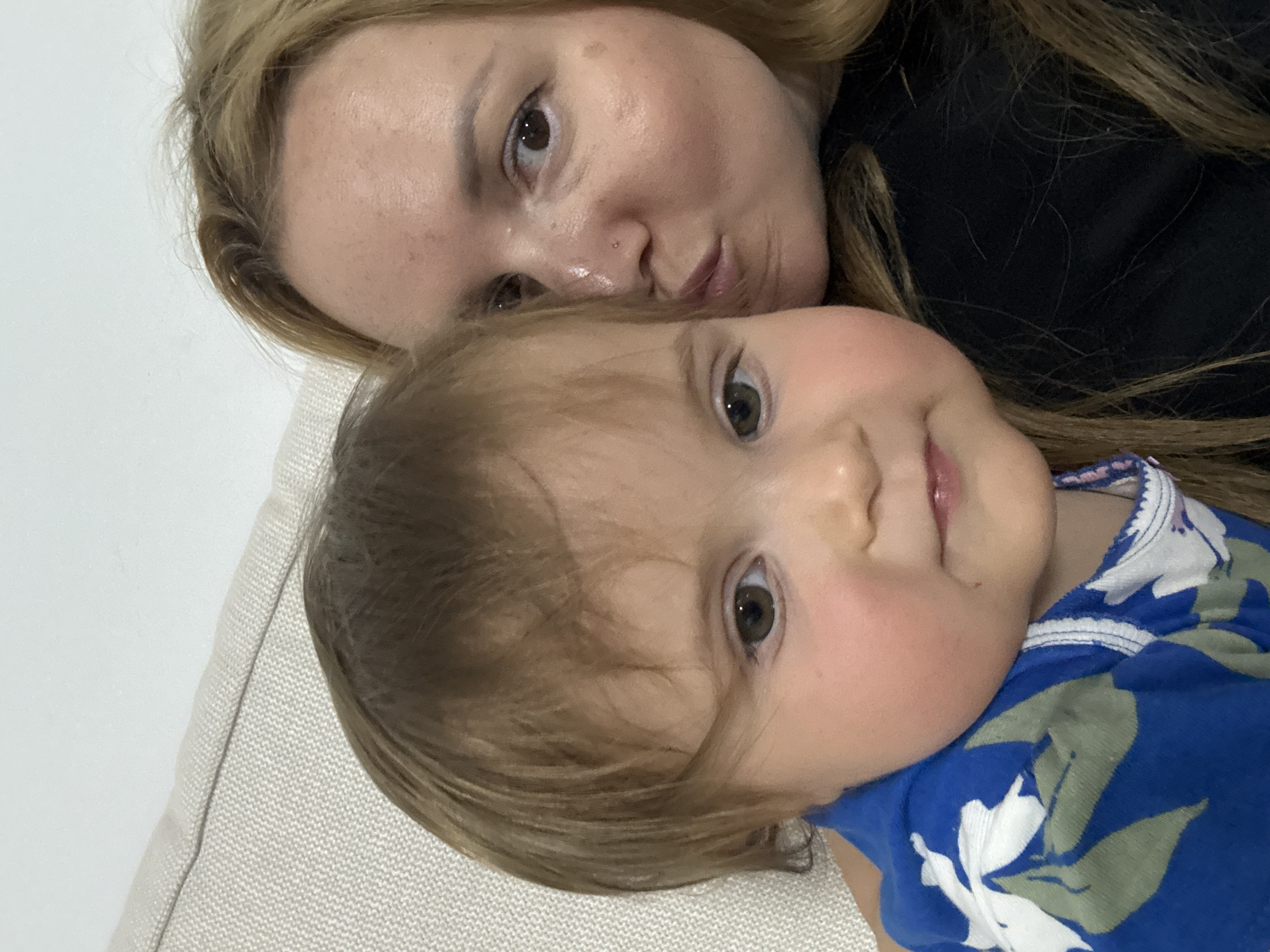 This indoor portrait, likely taken in Ramat Gan, Israel, features a woman and a baby lying closely together, looking towards the camera. The image is oriented such that the subjects appear rotated clockwise, with the woman positioned above and slightly behind the baby.
The woman has blond hair, fair skin with some subtle freckles, and light-colored eyes. She wears a dark top and has a small piercing on her nose. Her lips are slightly pursed in a playful or pouting expression.
Below her is a baby or young toddler with light brown hair, fair skin, and noticeable rosy cheeks. The baby has large, dark brown eyes and small, slightly parted lips, conveying a neutral and curious expression. They are wearing a vibrant blue top adorned with a white and green tropical floral pattern.
Both individuals are resting on what appears to be light-colored, textured upholstery, possibly a couch or bed, with a plain light-colored background behind them. The lighting is soft and even, suggesting an indoor setting during the daytime. The image captures an intimate and affectionate casual moment between the two subjects. No visible text is present in the media file.