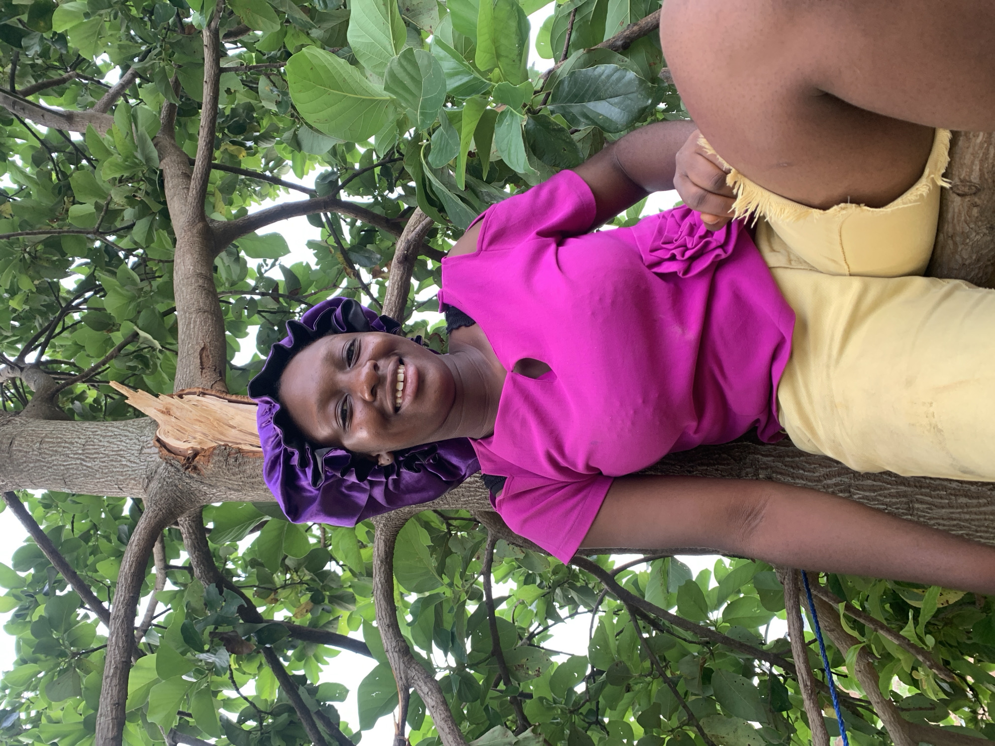 A medium shot depicts a woman perched in a tree in Agban, Nigeria. She wears a magenta top, yellow pants, and a purple bonnet. She is positioned on a tree branch, looking toward the camera with a smile. The surrounding tree branches and leaves frame the shot.