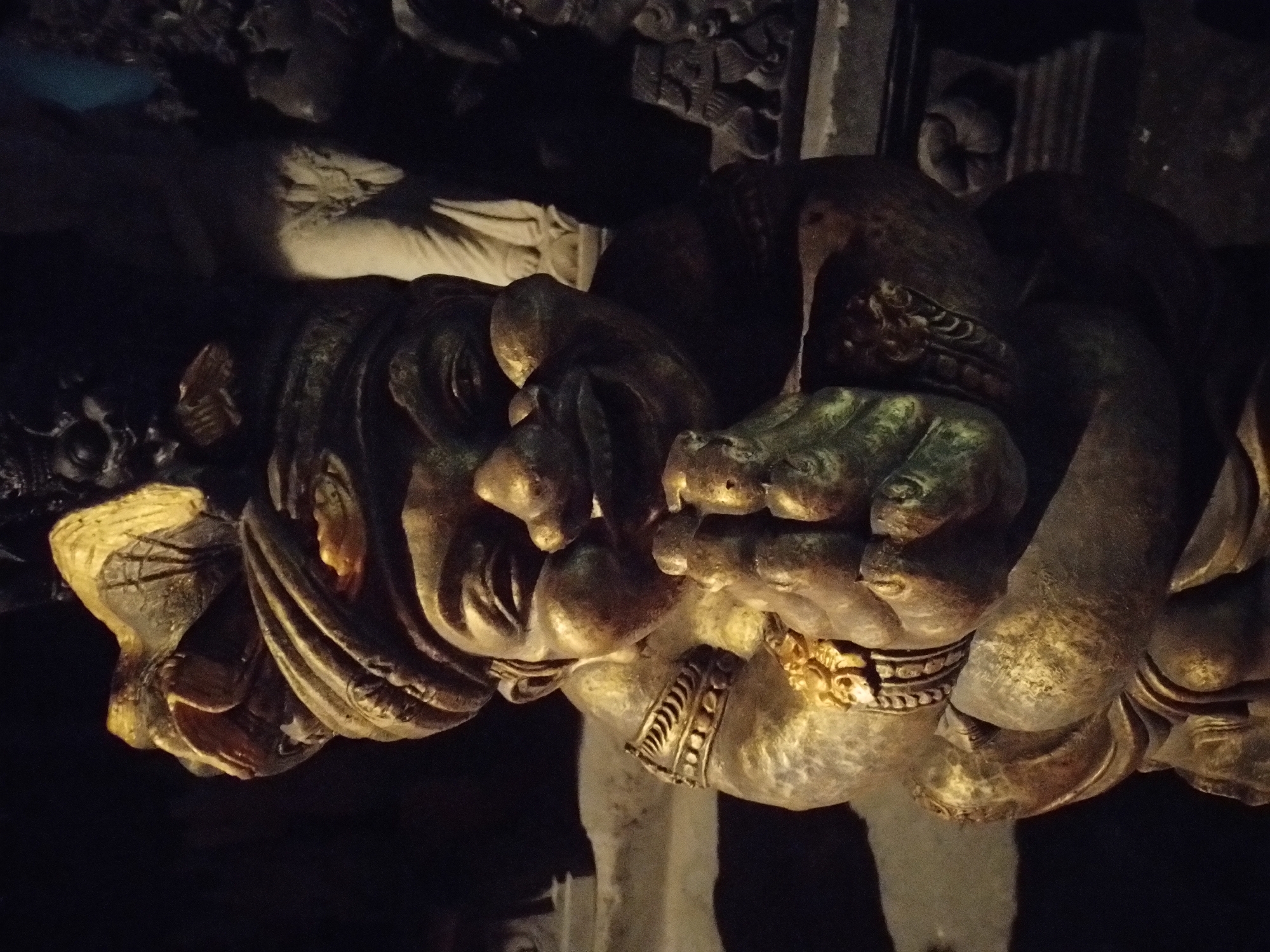 The media file showcases a close-up, dramatic shot of a large, intricately carved and gilded sculpture, likely a significant architectural detail or a standalone artwork. The primary subject is a mythical creature, characteristic of traditional Balinese art. Its stylized face dominates the foreground, featuring a wide, expressive mouth, possibly fangs, and ornate details around the eyes, though specific features are softened by the dark lighting.
The sculpture appears to be made of a dark material, possibly aged wood or stone, extensively adorned with gold leaf or paint that highlights its intricate carvings, textures, and contours. Hands or paws of the creature are visible, one of which appears to be clutching a smaller, bird-like figure or a complex decorative element. Embellishments resembling ornate bracelets or armbands are also visible on the limbs.
The scene is dimly lit, with focused artificial light dramatically illuminating the gilded portions of the sculpture, creating strong contrasts between the gleaming gold and deep, dark shadows. This lighting gives the piece a profound, majestic, and somewhat mysterious atmosphere. The background is largely indistinct and dark, with subtle hints of other carved elements or architectural structures in the distance. No specific activity is depicted as it is a static artwork. Given the context of Sukawati, Indonesia, the style strongly suggests this is a guardian figure or decorative element from a Balinese temple, palace, or traditional cultural building. No visible text is present in the image.