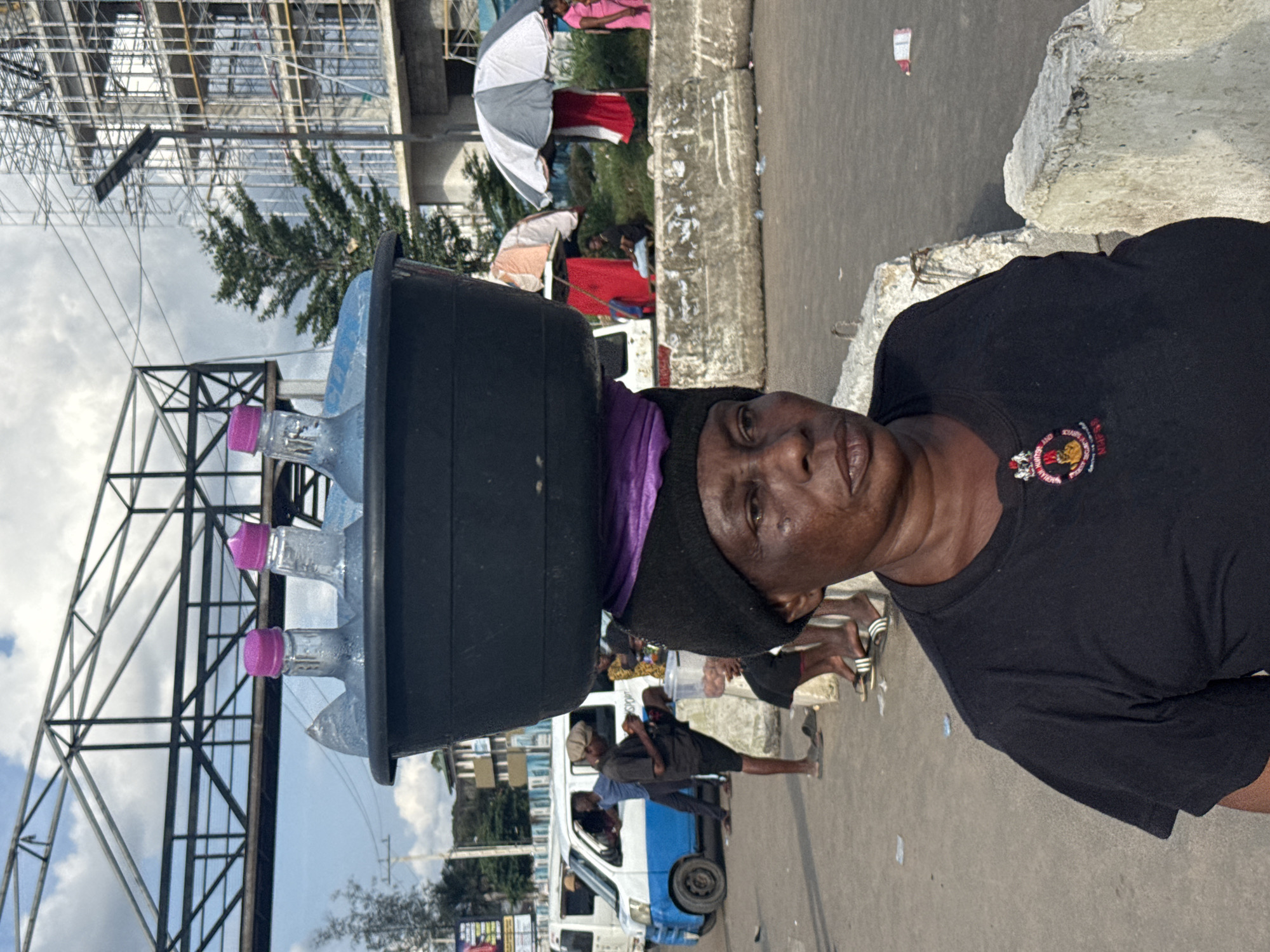 A woman, likely in her 50s or 60s, with dark skin and short, dark hair under a black beanie and a purple head wrap, is the primary subject of the image. She is wearing a black t-shirt with a circular emblem on the left chest. She is carrying a large black basin filled with bottled water on her head. The bottles have pink caps. The scene appears to be a street or marketplace in Owerri, Nigeria. In the background, there is a blue and white vehicle, construction scaffolding on a multi-story building, a pedestrian bridge, and other people standing or sitting. The ground is paved, and there are concrete barriers visible. The sky is cloudy with patches of blue. The woman is looking slightly to her right.