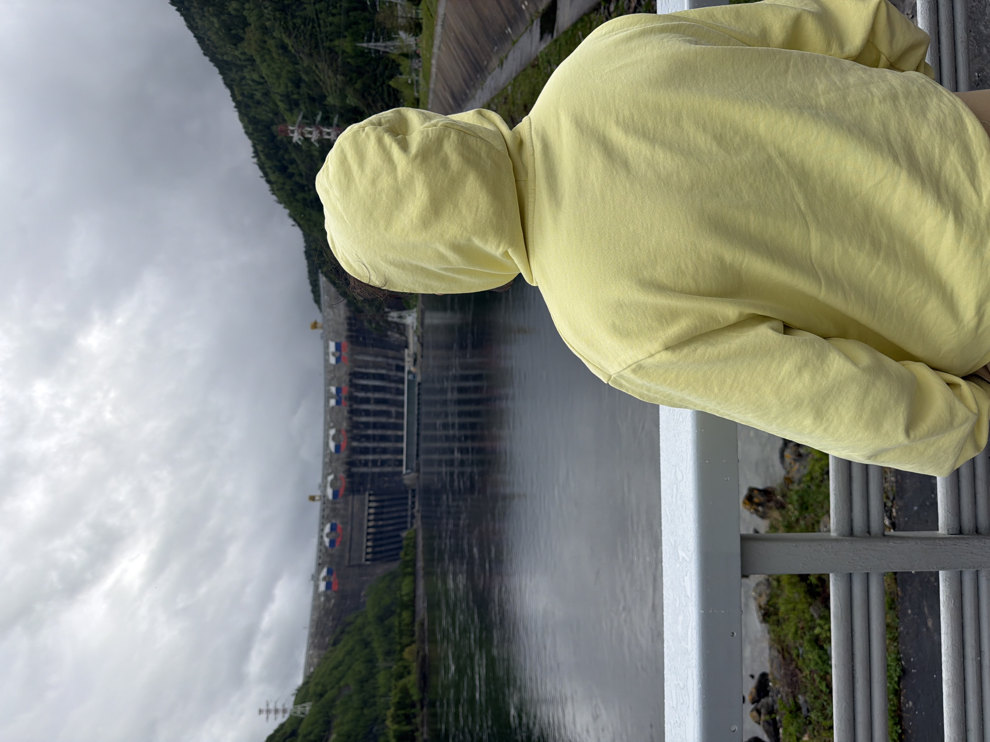 A person wearing a yellow hoodie is seen from behind, leaning on a gray metal railing and looking out at the Sayano-Shushenskaya Dam on the Yenisei River in the city of Cheremushki, Russia. The dam is a large concrete structure with a series of gates. The water of the river fills the frame between the dam and the railing. Green trees line the banks of the river. The sky is overcast and gray. Transmission towers are also visible along the mountain landscape.
