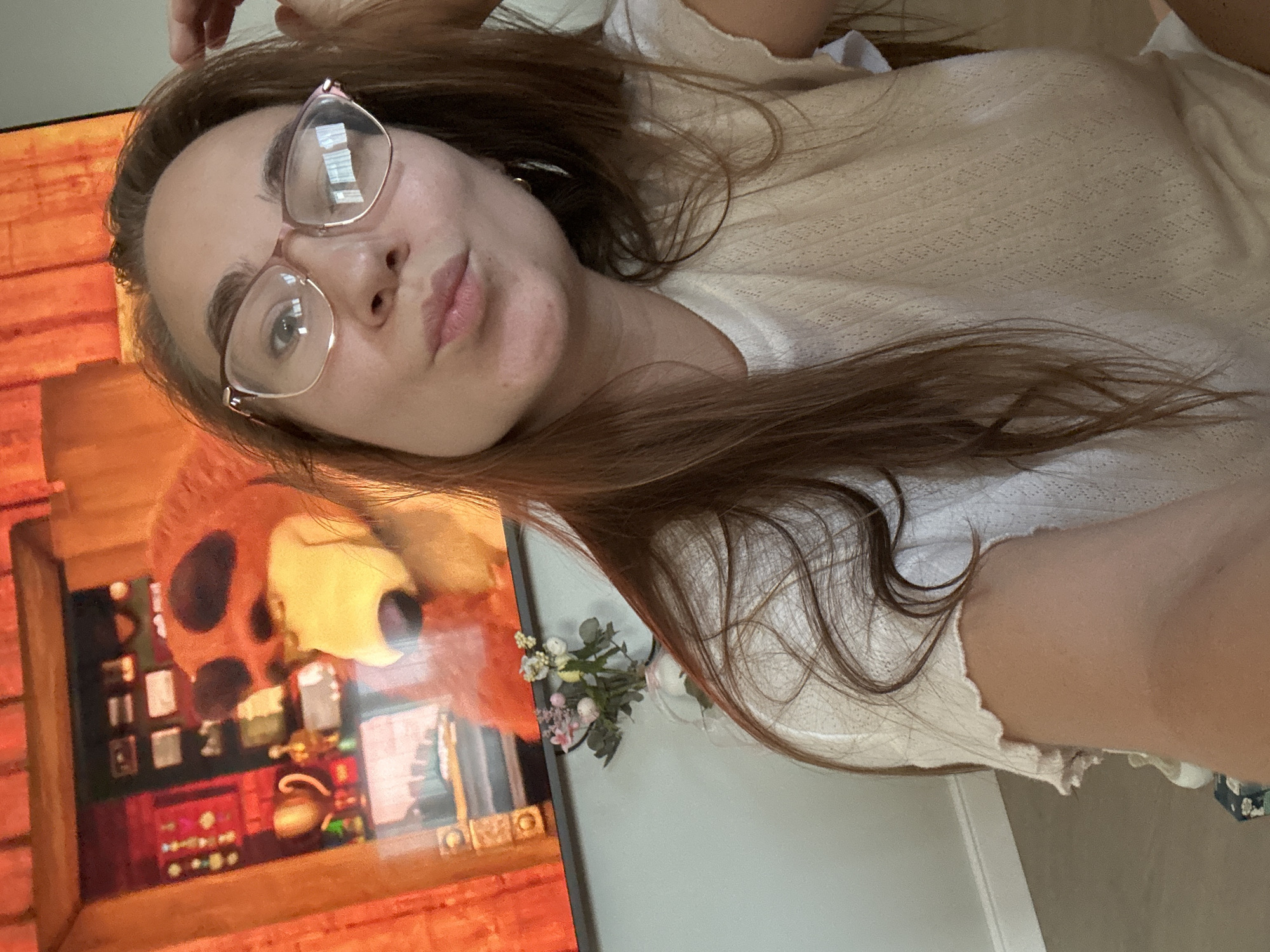 A close-up, slightly tilted selfie captures a young woman with long, reddish-brown hair, lying down in what appears to be an indoor setting in Kharnavy, Russia. She is wearing light-framed, clear glasses and a light-colored, possibly cream or off-white, textured top. Her expression is a playful pout or "duck face," conveying a casual and relaxed mood. Her left hand is partially visible at the top of the frame, gently touching her hair.
To the left of the woman, a large television screen is visible, displaying an animated scene. The main character on screen is a furry, orange or reddish-brown creature, possibly a lion or bear, with large eyes and a prominent snout. The setting within the animation appears to be a detailed, warm-toned indoor room, featuring shelves, various small objects, and a teapot. Next to the television, a small bouquet of white and pink artificial flowers with green leaves is placed against a light-colored wall. The overall ambiance suggests a comfortable and informal moment. No visible text is present in the image.