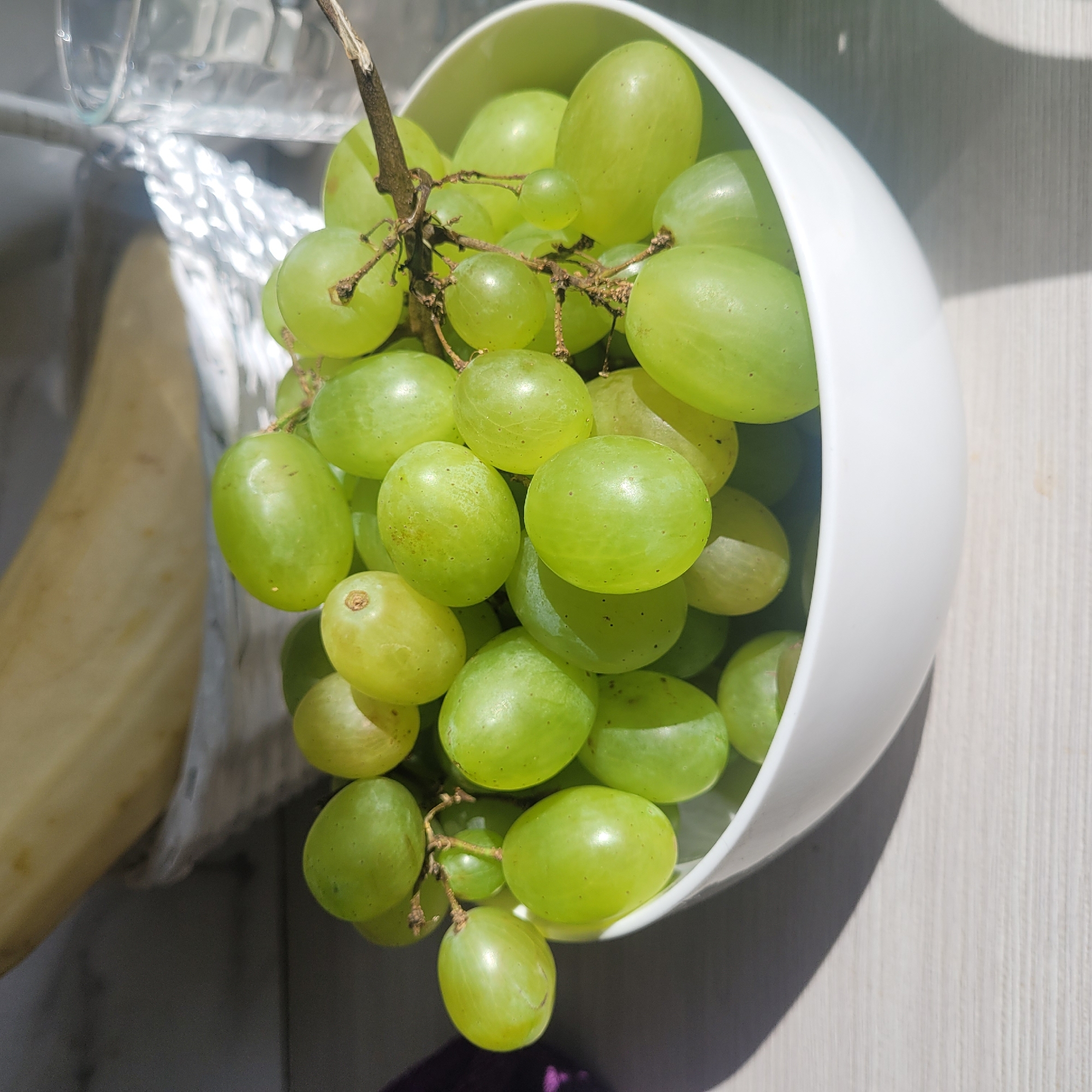 A white bowl filled with green grapes sits on a light-colored, horizontally-grained surface. The bowl is positioned slightly off-center to the right, with the grapes spilling over the rim and a few strands of the bunch extending beyond. The grapes are round to oval in shape, with some exhibiting slight variations in color from light green to a more yellowish-green. The surface they are on casts a distinct shadow to the left, suggesting a light source from the right. In the background, out of focus, is a white woven basket containing a pale yellow, elongated object, possibly a banana, and a clear glass with ice cubes. The overall scene is brightly lit, with harsh shadows indicating direct sunlight. There are no people or animals present in the image. The scene does not contain any identifiable landmarks or specific elements that would indicate a location within Jūrmala, Latvia.