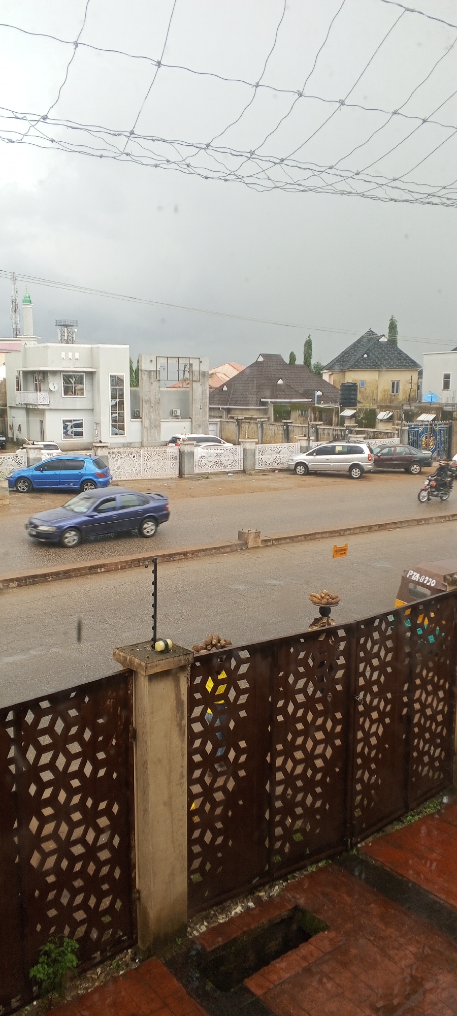 The image captures a street scene in Jos, Nigeria, under an overcast sky, suggesting a potential for rain. In the foreground, a dark brown metal gate with an intricate geometric pattern is visible, with a concrete pillar supporting it. Beyond the gate, a paved road is wet, indicating recent or ongoing precipitation. Several vehicles are present: a blue sedan and a darker blue sedan are moving on the road, a silver SUV is parked on the side, and a maroon sedan is also on the road. A person is riding a motorcycle in the distance. Further back, various buildings line the street, including modern structures and more traditional homes with distinct roof designs. One building features a minaret, suggesting the presence of a mosque. A person is seen on the right side of the road carrying a tray of goods on their head, possibly a street vendor. The overall impression is a typical urban environment in Jos, Nigeria, with everyday activities taking place despite the weather conditions.