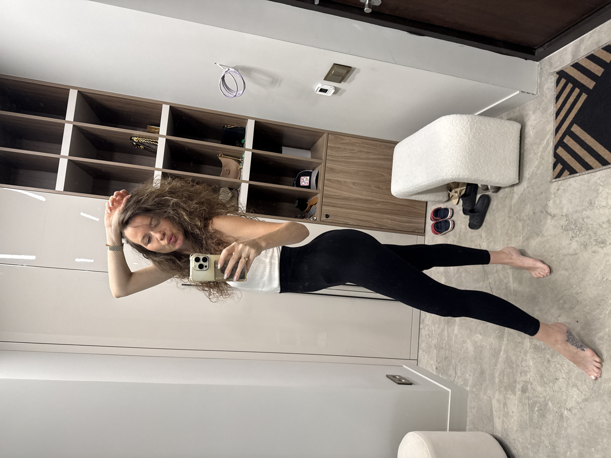 A young woman with curly brown hair and dark leggings is taking a mirror selfie in what appears to be a dressing room or hallway. She is wearing a white top and has her right arm raised, touching her hair. She is puckering her lips as if blowing a kiss. To her right is a tall wooden shelving unit filled with various bags and a hat. In the foreground, there is a white upholstered bench and several pairs of shoes, including red and blue flip-flops and black Crocs. The floor is tiled with a gray, marble-like pattern. A dark wooden door is visible on the far right. The image does not contain any specific visual cues to definitively place it in Khimki, Russia.