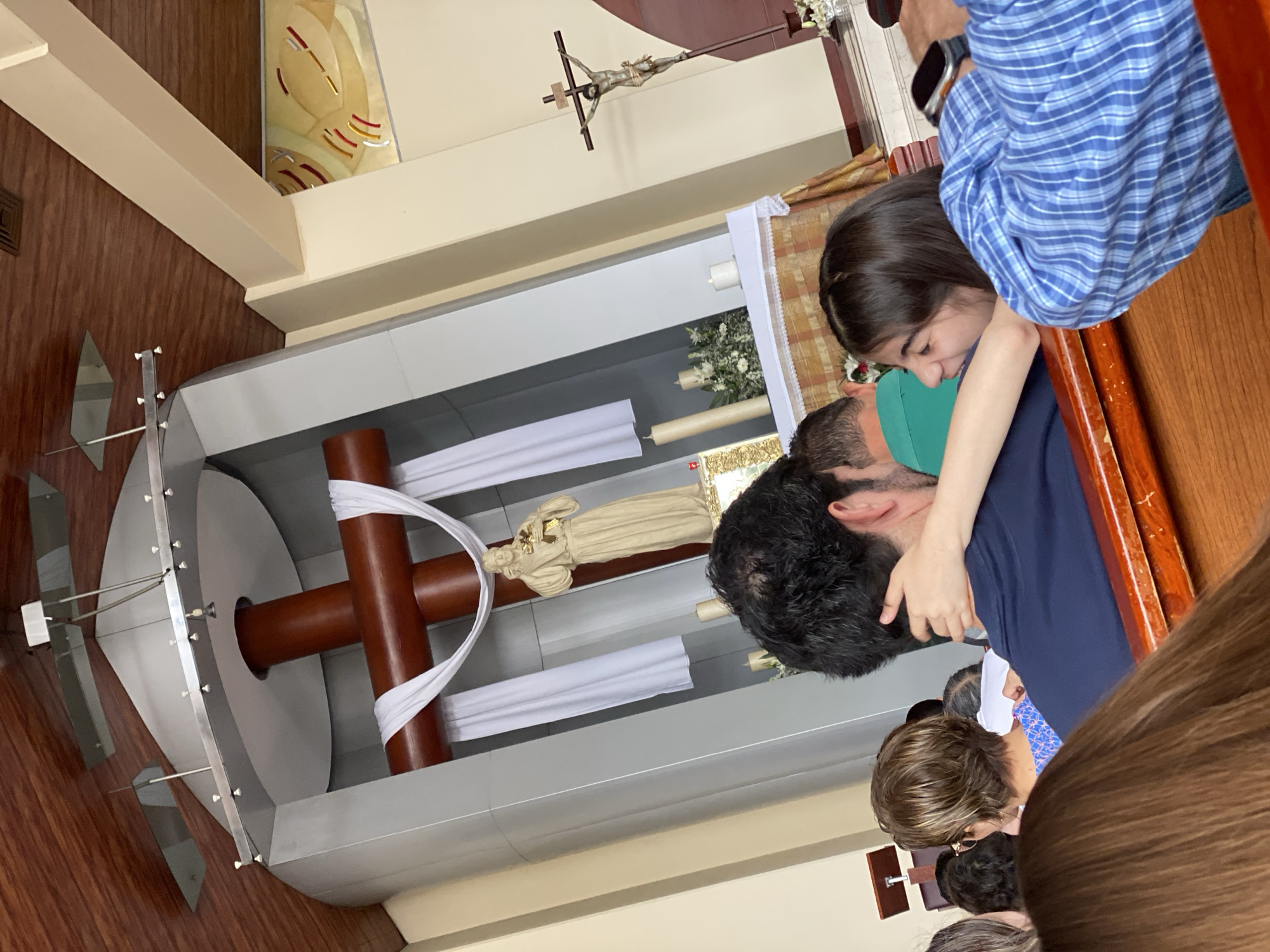 The image shows the interior of what appears to be a church in San Bernardino Tlaxcalancingo, Mexico. A wooden cross is visible, draped with a white cloth. There is a statue, likely of a saint or religious figure. People are present, with some appearing to interact and embrace. The specific details are difficult to discern given the angle and partial obstruction of the view.