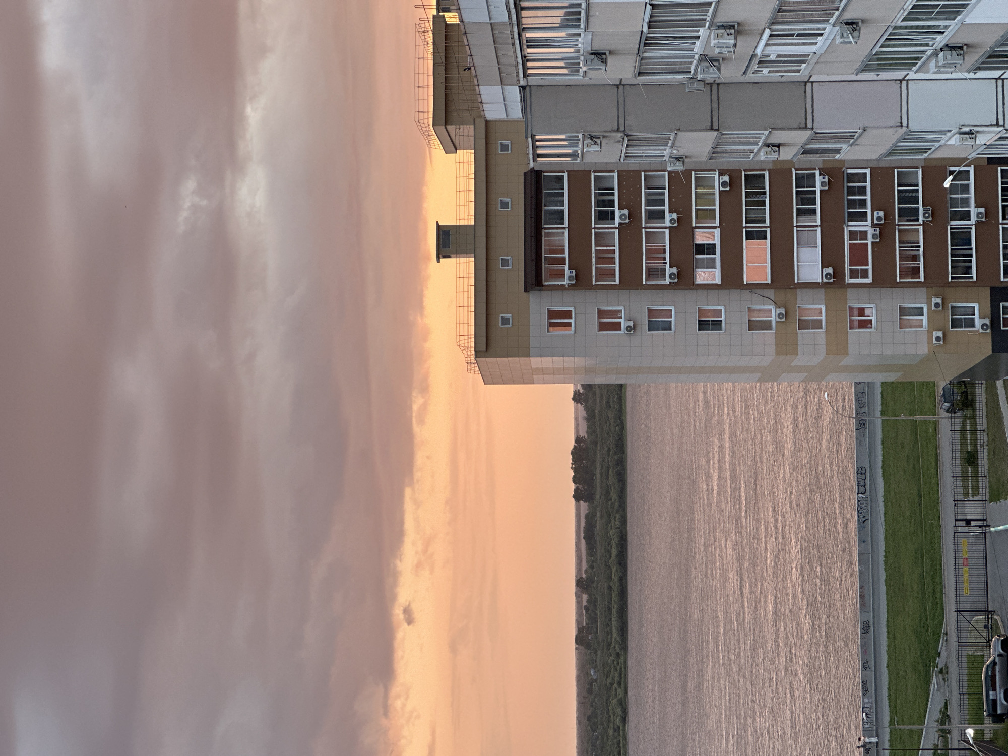 The image depicts a view of the Amur River and apartment buildings in Khabarovsk, Russia, at sunset. The sky is filled with warm-toned clouds in shades of orange, pink, and gray. The Amur River occupies the lower left and center of the frame, its surface reflecting the colors of the sky. On the far bank of the river, a line of trees and vegetation is visible. In the foreground, several apartment buildings are prominent. The closest building on the right is light gray and white, with numerous windows and air conditioning units. To its left, another apartment building features a facade of tan and brown sections, also with many windows and air conditioners. The top of the buildings shows railing and rooftop structures. Along the riverbank, a promenade or walkway is visible, with graffiti on a low wall. A strip of green grass and a fence separate the promenade from a road below, where two vehicles are parked. The scene suggests a residential area of Khabarovsk along the Amur River.