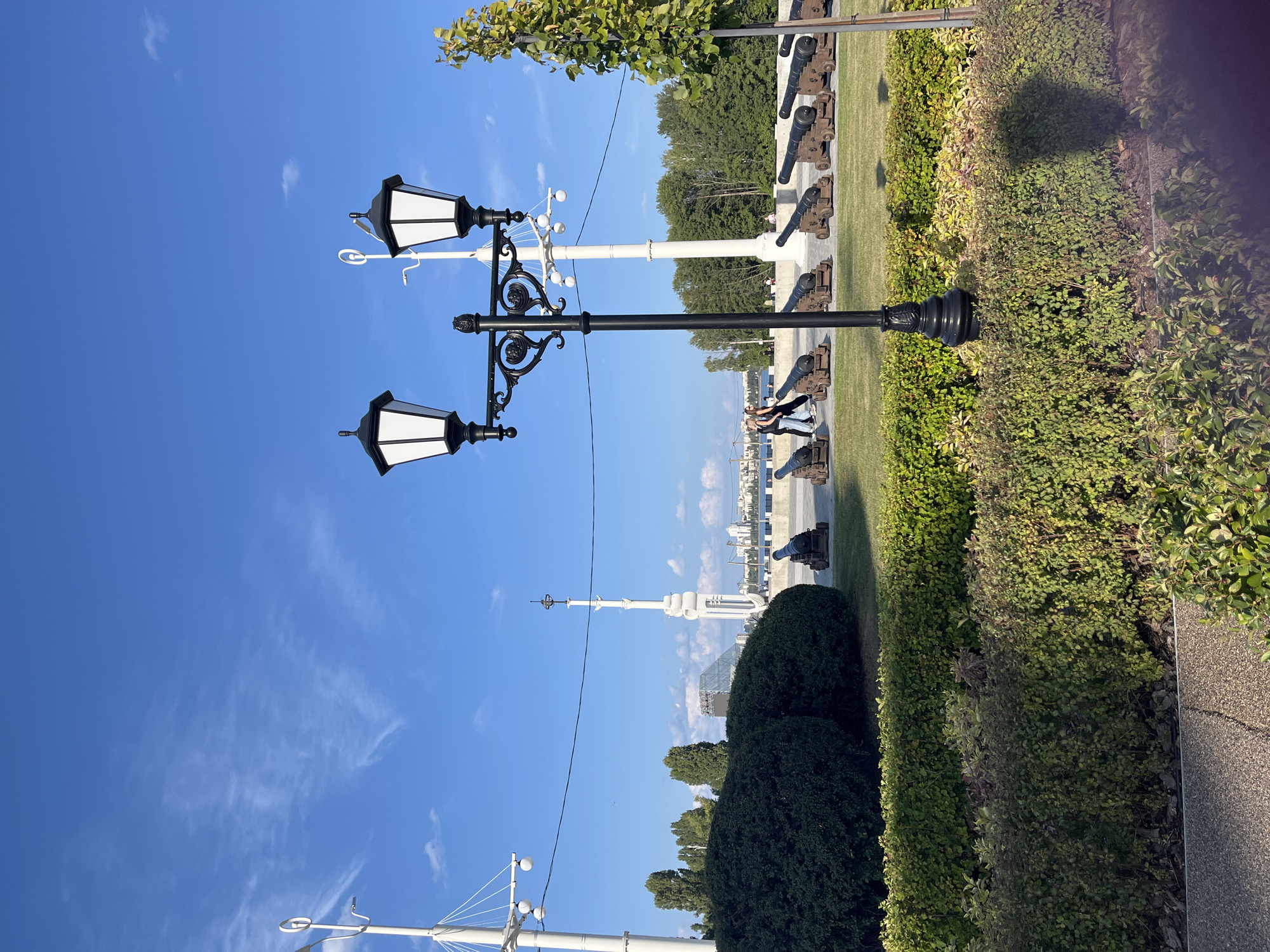 The image captures a sunny day with a clear blue sky and scattered clouds. In the foreground, a black ornate lamppost with two lamps stands prominently. Behind it, a neatly manicured green hedge forms a boundary. Further back, a line of dark-colored cannons with wooden wheels is arranged on a paved surface. Two individuals, a man and a woman, are walking together amongst the cannons. A large, dark green, rounded hedge dominates the left side of the middle ground. Beyond the hedge and cannons, a body of water is visible, with a city skyline in the distance featuring various buildings and a distinctive white tower. Tall trees line the horizon. The setting appears to be an outdoor public space, possibly a park or embankment, given the presence of historical artifacts, greenery, and a waterfront view. The location is identified as Novaya Usman, Russia.