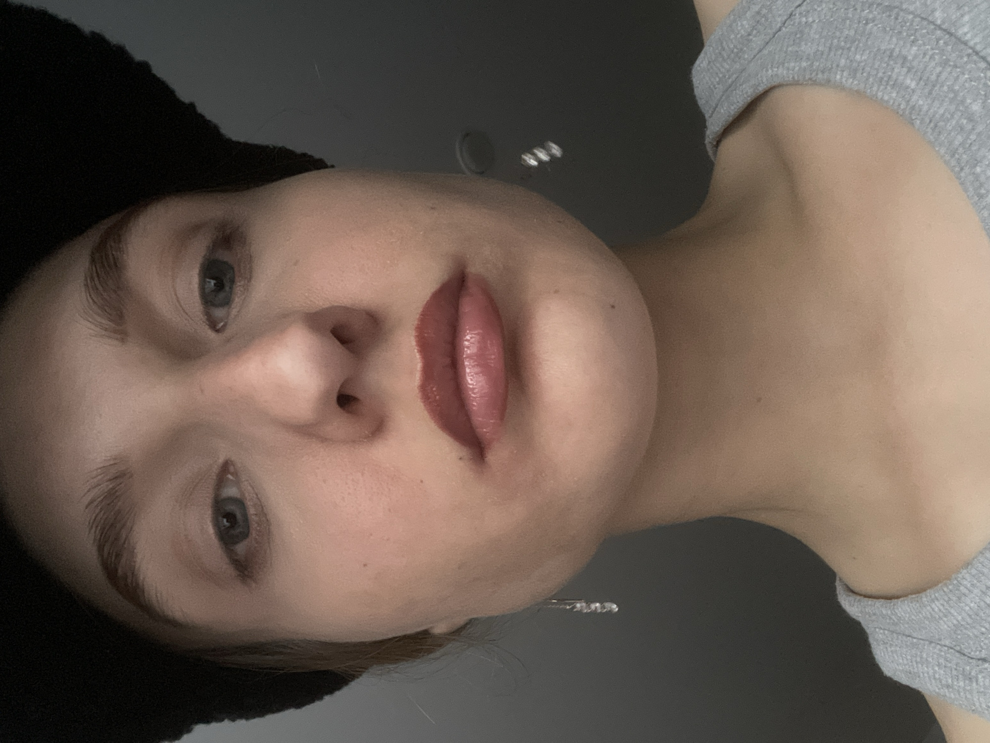 A close-up, low-angle shot of a woman with gray eyes and glossy lips. She is wearing a gray tank top. The background is dark gray. The location is not discernible in this image.