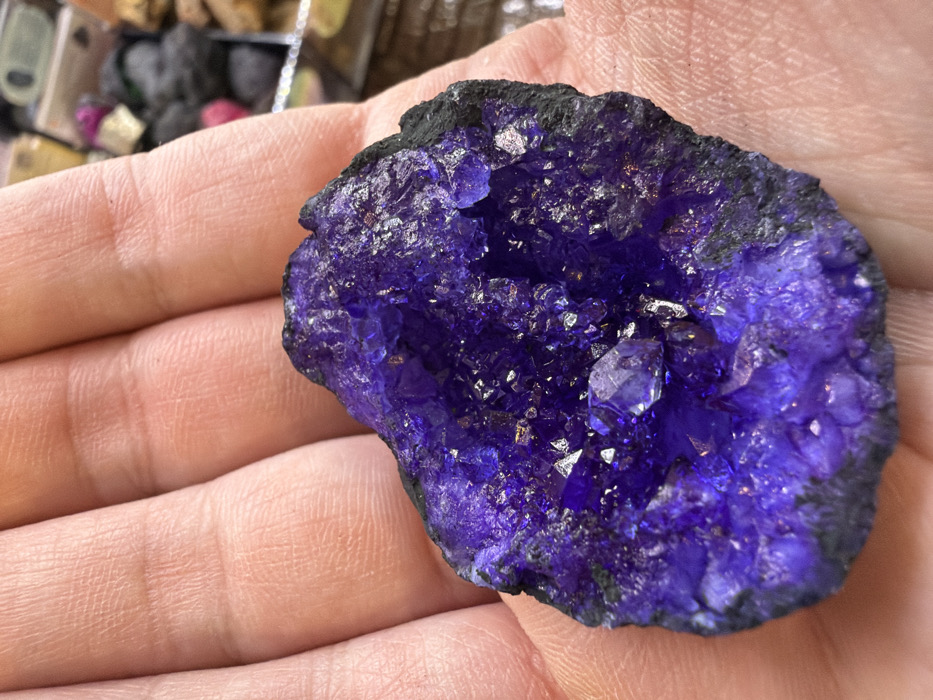 An adult human hand, exhibiting light skin tone and visible palm and finger creases, holds a roughly palm-sized, irregular purple mineral geode in an open-palm presentation. The geode features a central cavity densely lined with small, reflective, deep purple crystalline structures. A few larger, faceted crystal points are observable within the cluster. The outer perimeter of the geode consists of a dark, rough rock matrix. The background is out of focus, showing indistinct forms and colors consistent with a collection or display of other mineral or stone objects. The scene is located in the city of Arucas, Spain.