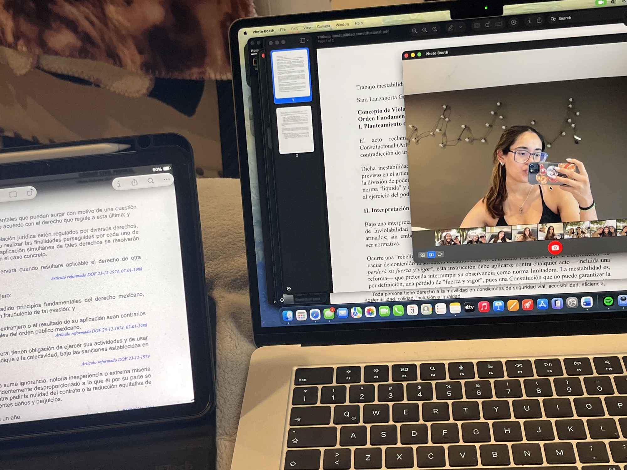 The image shows a desk setup with a laptop and a tablet. The laptop displays a document titled "Trabajo inestabilidad constitucional.pdf" and a Photo Booth application showing a young woman with glasses taking a selfie with a phone. The tablet displays a document in Spanish, possibly related to law. The environment is blurry and out of focus, but appears to be indoors. There is no indication of a specific location in San Bernardino Tlaxcalancingo, Mexico.