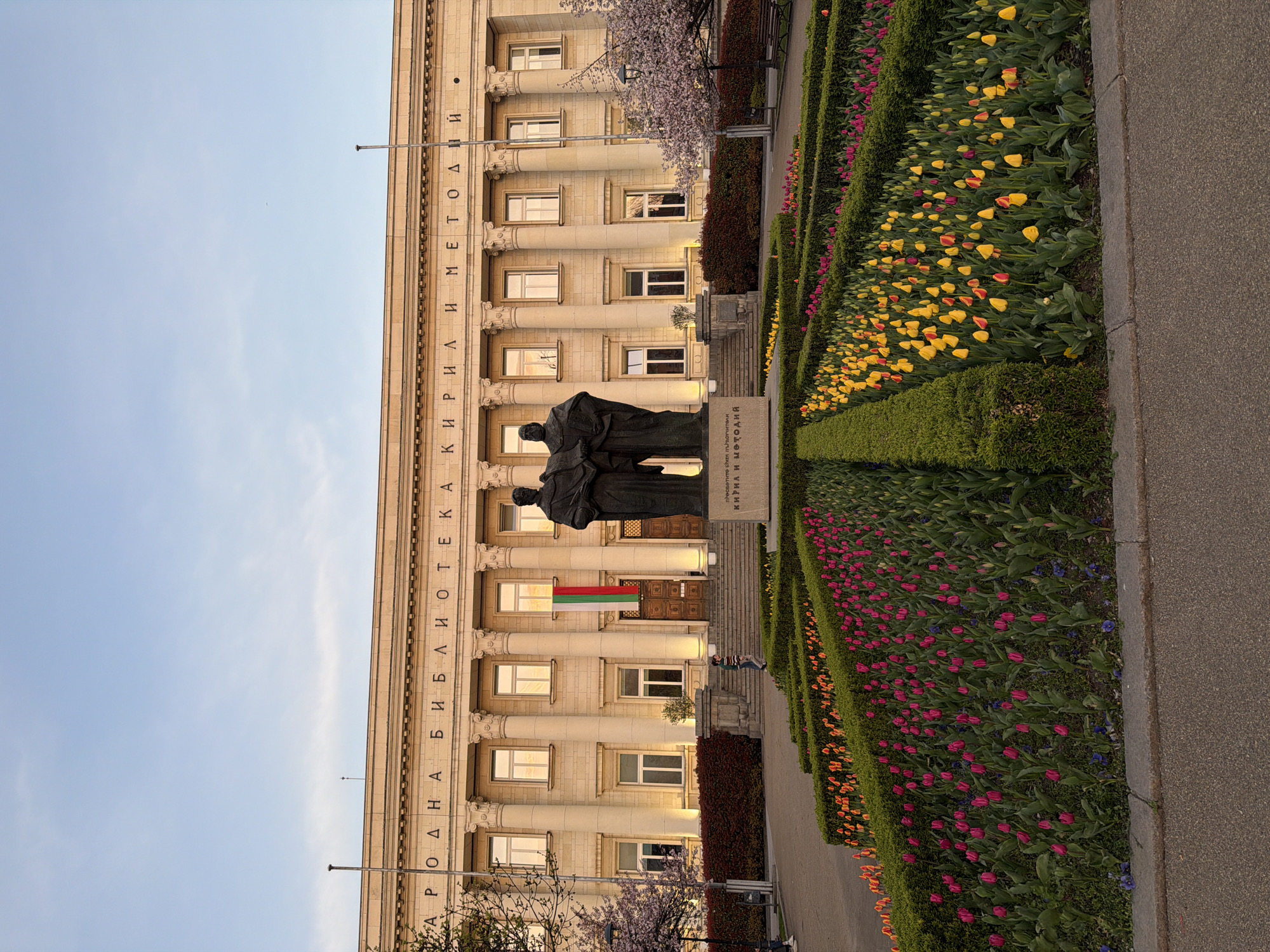 The image captures the National Library "St. Cyril and St. Methodius" in Sofia, Bulgaria, viewed from ground level. The library's facade is visible, featuring large columns, windows, and inscribed Cyrillic text. In front of the building stands a monument depicting two figures, likely Cyril and Methodius. The Bulgarian flag is displayed above the building's entrance. Flower beds planted with tulips in various colors are arranged along the walkway leading to the building. A paved pathway borders the flower beds. The sky above the building is light blue, indicating either early morning or late afternoon.