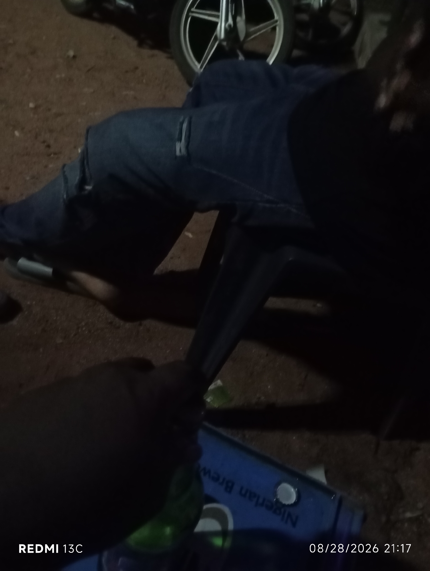 The image shows a person's legs in blue jeans, seated on a dark chair. The person is wearing a dark shoe or sandal on their left foot. In the foreground, a hand is holding a green glass bottle with a blue label that reads "Nigerian Brew". The background is dark and appears to be an outdoor setting with loose soil and a motorcycle wheel visible. The timestamp "08/28/2026 21:17" is superimposed on the bottom right of the image, and "REDMI 13C" is on the bottom left, indicating the device used for capture. The scene is consistent with an informal gathering in Shendam, Nigeria, possibly at night, given the lighting conditions and timestamp.