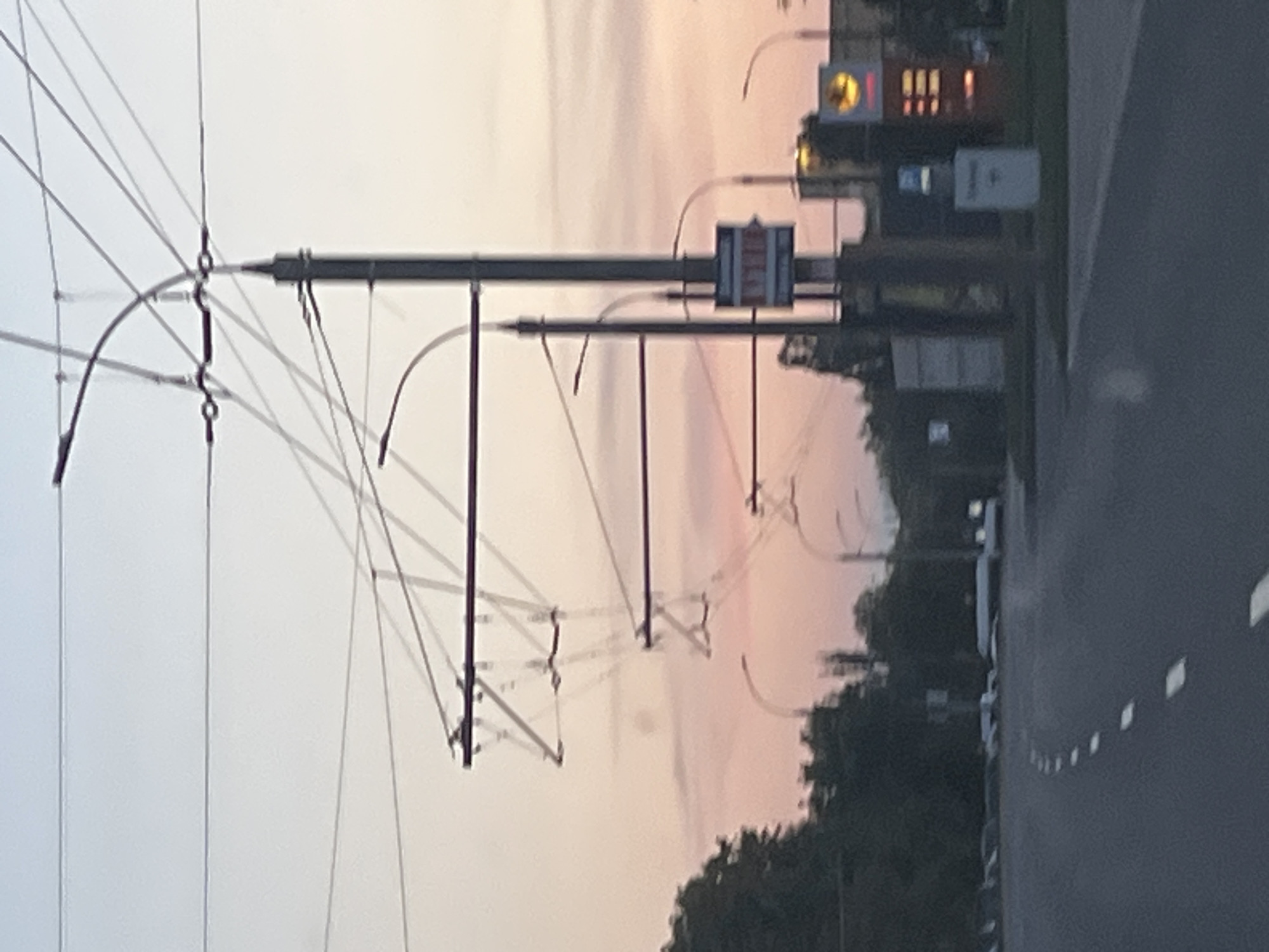 The image depicts an urban scene, possibly at dusk or dawn, characterized by a network of overhead wires and trolleybus poles against a soft, pastel-colored sky. The foreground shows a road with a dashed white line marking its center. On the right side of the road, there are buildings with illuminated signs, including what appears to be a gas station with a yellow circular logo and a red display showing prices. A street sign with red and white markings is visible on a pole. Trees line the left side of the road. The overall impression is of a street in a city, with infrastructure for public transportation, such as trolleybuses, evident. This scene is consistent with observations from the city of Brest, Belarus.