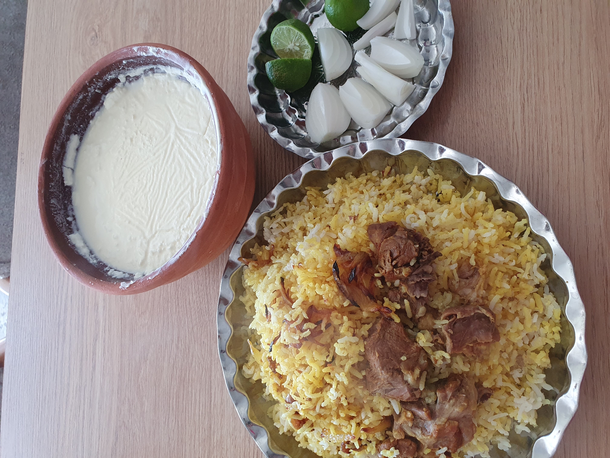A top-down shot captures a meal of biryani, yogurt, and sliced onions with lime wedges, presented on a wooden table. The biryani, a savory rice dish with tender meat and fried onions, is served in a silver-colored, scalloped-edged platter. To the right, a small silver platter holds wedges of white onion and lime. A rustic, terracotta bowl filled with thick, white yogurt sits at the top right of the frame. The wooden table's grain is visible, providing a warm backdrop to the food.