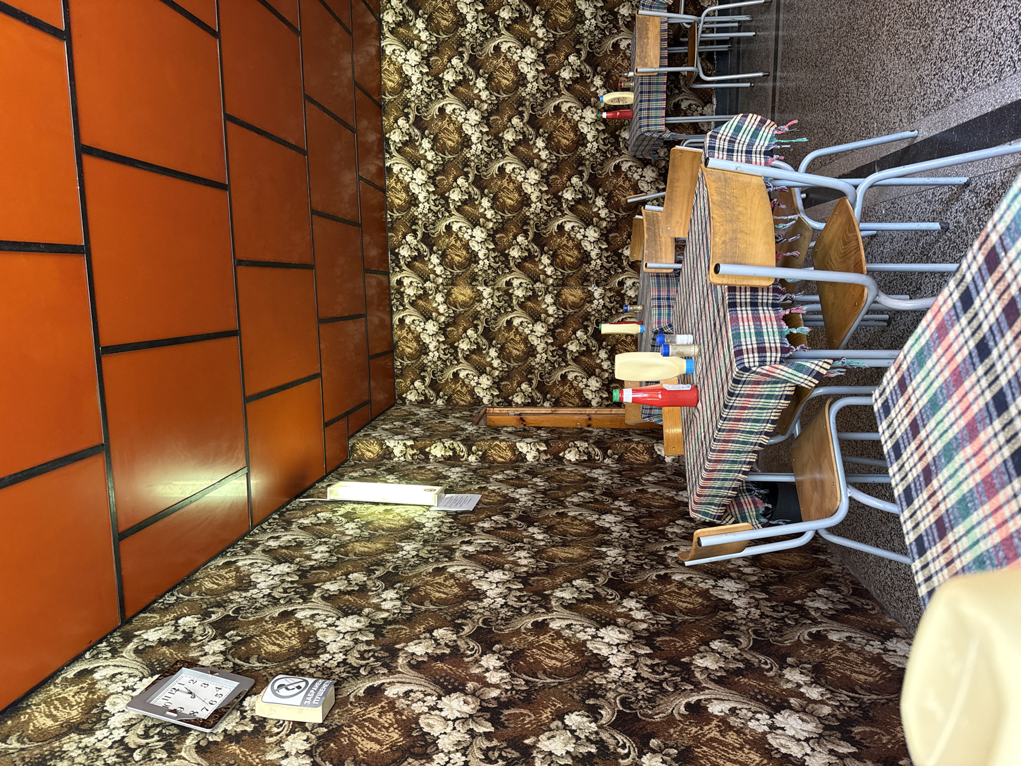 The image depicts an interior space in Elhovo, Bulgaria, characterized by a distinctive aesthetic. The walls feature a combination of orange tiled paneling and patterned wallpaper. The floor is covered in floral patterned carpeting. There are objects, including a clock and a small sign (likely in Bulgarian) resting on the floor near the orange wall. The room is furnished with tables and chairs. The chairs are stacked and draped with plaid fabric covers. Food items, including a bottle of ketchup are visible on the tables. The layout includes a raised platform with the same patterned wallpaper and carpeting. The room has an unusual orientation, with the camera position tilting the perspective.