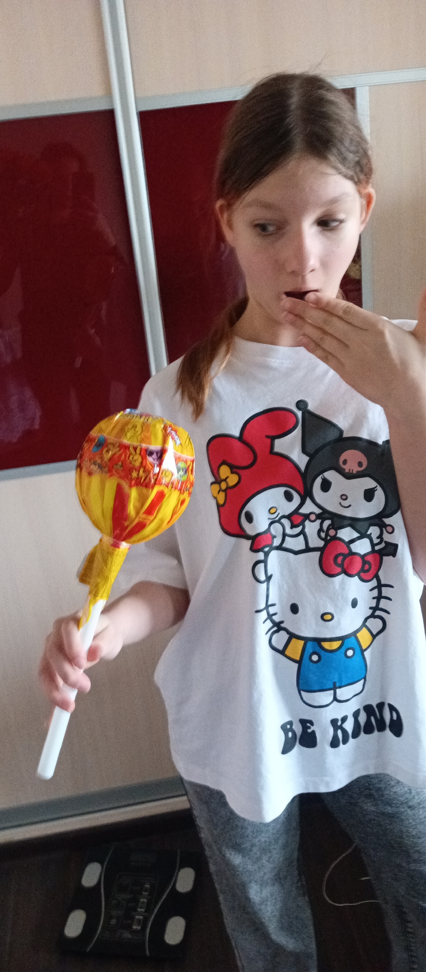 A young person with light brown hair in a ponytail is holding a large lollipop with a yellow and red striped wrapping in their left hand. They are wearing a white t-shirt with a cartoon graphic featuring Hello Kitty, My Melody, and Kuromi, and the phrase "BE KIND" printed below. The person's right hand is partially raised to their mouth, and they are looking towards the lollipop. The background includes a red cabinet with white trim, and a dark floor with a visible scale. The location is indoors. The city and specific location are Unknown.