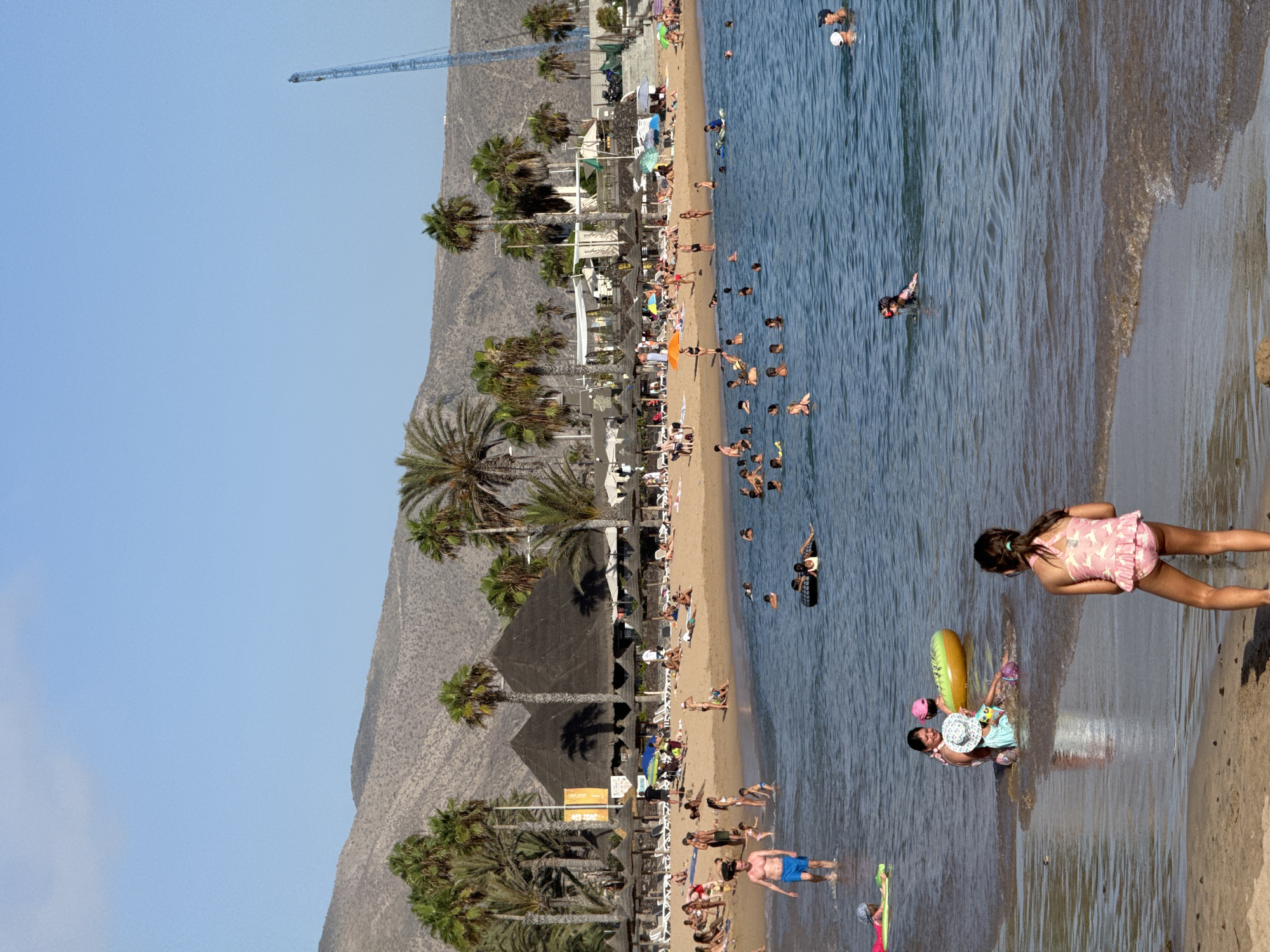 This is a photograph of a crowded beach scene in the city of Arona, Spain. The image is taken from a low angle, looking towards the water and the beach.
In the foreground, the sandy shore is visible, with a young girl in a pink swimsuit standing facing away from the camera. Further out in the water, several people are swimming and playing. A group of people, including a woman and two children, are in the shallows near the shore, with one child sitting on an inflatable ring.
The middle ground of the image shows the ocean teeming with swimmers. A man in blue swim trunks is walking in the water, and several other individuals are scattered throughout the water, some with inflatable toys. In the distance, the beach is lined with umbrellas and lounge chairs, with many people sunbathing and relaxing. Palm trees are scattered across the beach and the background, with a large mountain range visible in the far distance. A tall construction crane is also visible in the upper right portion of the image, indicating ongoing development in the area. The sky is clear and blue, suggesting a sunny day.