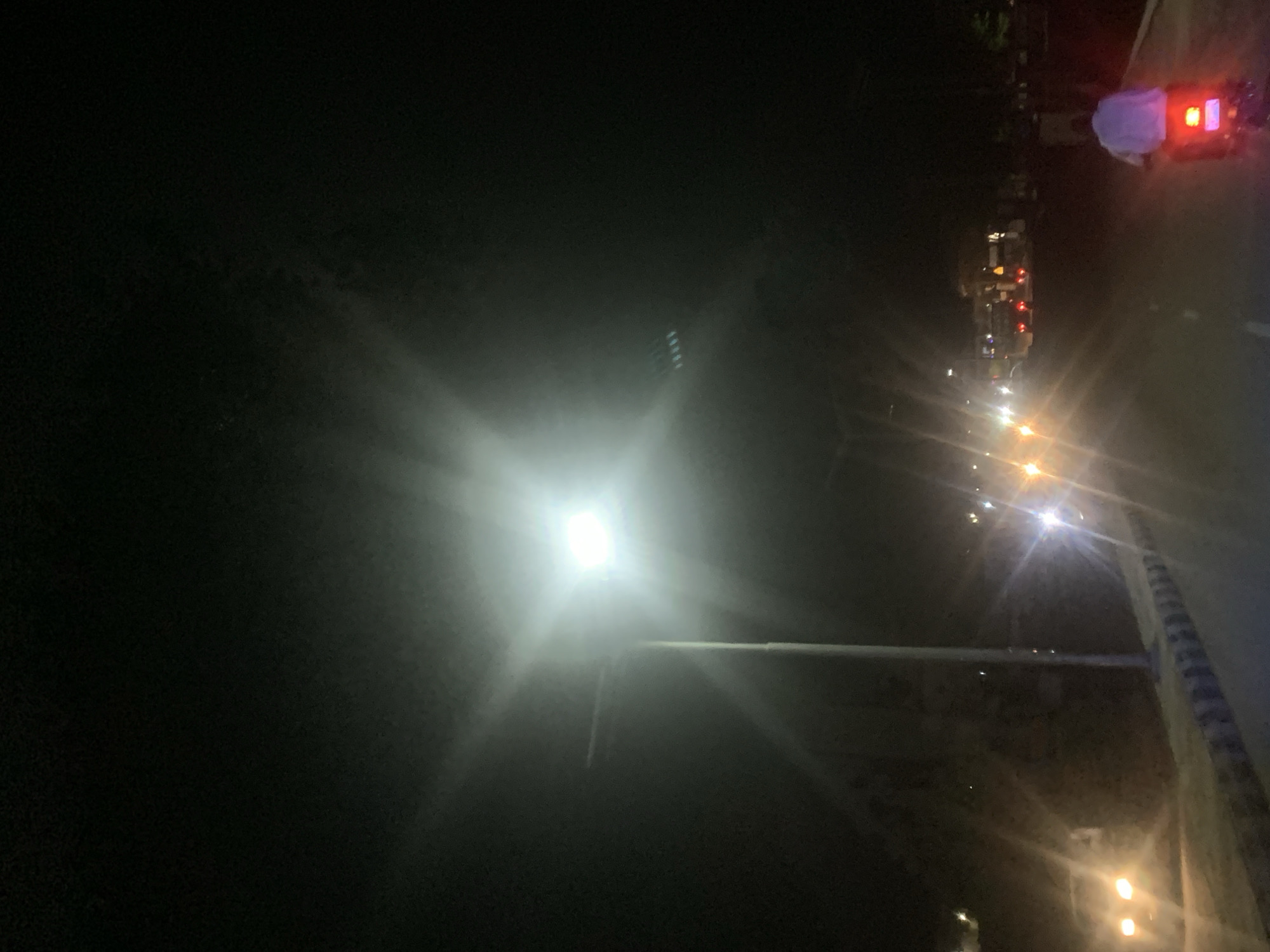 The image depicts a night scene in Adankolo, Nigeria. A bright street light dominates the center of the frame, casting a strong glare and rays of light. To the right, a line of other street lights recedes into the distance. A few vehicles with red lights are visible on the road. One person wearing a white top, possibly near a vehicle, is seen at the top-right of the image. The left side of the image is mostly dark, suggesting a less illuminated area. A concrete or metal structure is visible on the right side of the image and leads to the middle bottom part of the image.