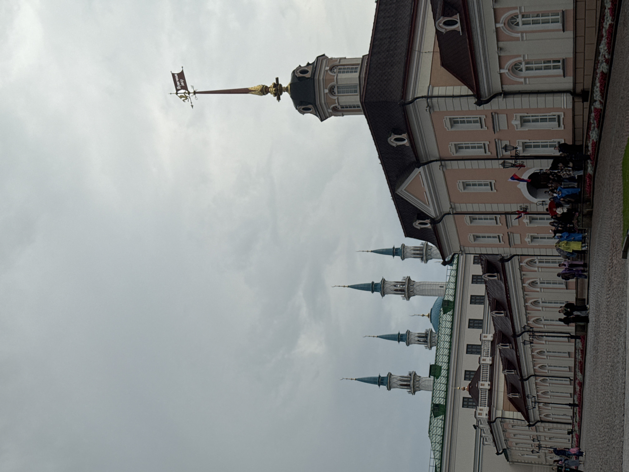 The image depicts the Kul Sharif Mosque in Kazan, Russia, a prominent landmark within the Kazan Kremlin. The sky is overcast, casting a muted light over the scene. In the foreground, a cobblestone plaza leads up to a collection of buildings. The main subject on the right is a pink and white multi-story building with a dark roof, featuring numerous arched windows and decorative elements. A tall, slender tower with a gold spire topped by a weather vane rises from the roofline. To the left, the iconic minarets of the Kul Sharif Mosque, with their blue conical tops and crescent moon finials, are visible against the cloudy sky. A mosque dome, also blue, is partially obscured by the buildings in the foreground. A group of people, some holding umbrellas and wearing colorful clothing, are gathered in front of the pink building. The overall impression is one of historical and architectural significance under a subdued sky.