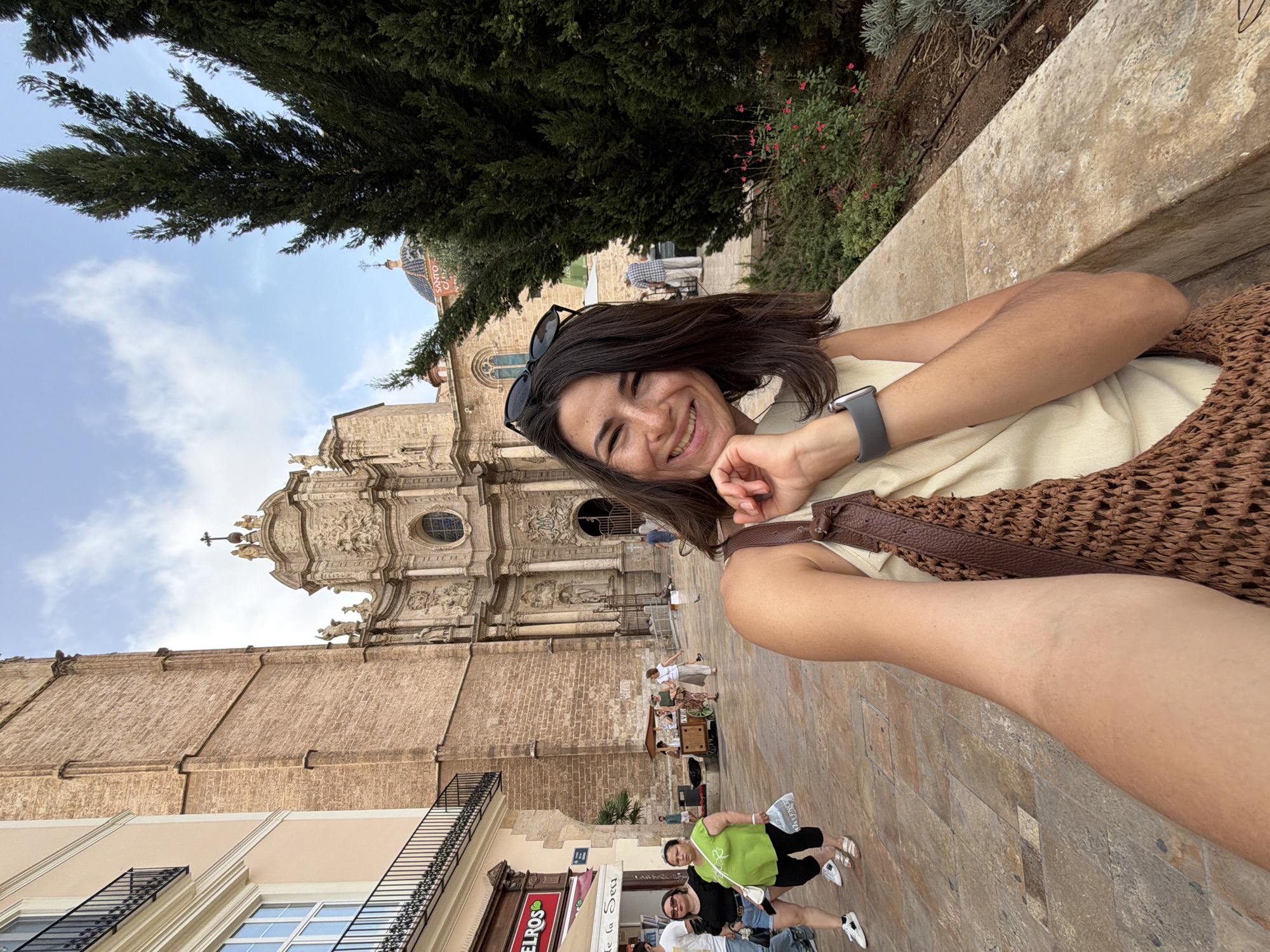 In this image, a woman is taking a selfie outdoors in an urban environment. She is smiling and leaning her head on her hand, with her arm visible in the foreground. She is wearing a light-colored top and a brown woven bag is slung over her shoulder. A smartwatch is on her left wrist. Behind her, a large stone building with intricate architectural details, possibly a cathedral, is visible. Several other people are walking in the background on a paved area, and there are trees and a cloudy sky. The scene appears to be a public square in a European city, given the architecture.