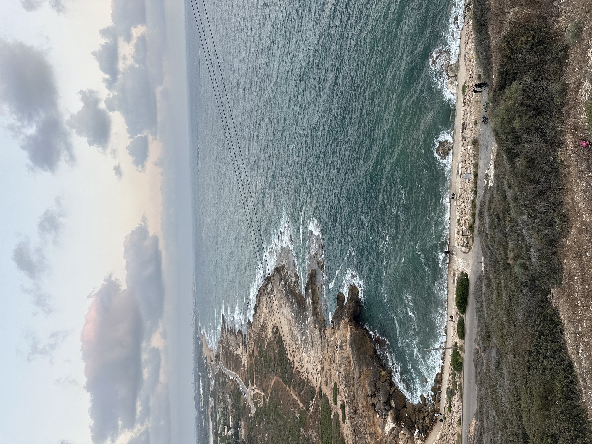 The image captures a scenic coastal view in the Mate Asher Regional Council, Israel, likely from an elevated vantage point. The foreground features dry, scrubby vegetation on a hillside sloping down towards a paved path and a rocky coastline. Several people are visible walking and sitting along the path, some near parked motorcycles.
The path runs parallel to the sea, bordering a rocky shore where waves crash against the stones, creating white foam. Beyond the immediate coastline, the sea stretches out, its surface rippled by the wind. The water is a deep teal color, transitioning to a lighter blue towards the horizon. In the distance, a faint line of buildings suggests a city or town along the coast.
The sky above is filled with a mix of clouds. Some clouds are a soft pinkish-white, possibly indicating either sunrise or sunset, while others are a more muted gray. Electrical wires are visible spanning across the upper right portion of the frame, suggesting some form of infrastructure in the area. The overall impression is of a natural coastal landscape with signs of human activity, possibly a popular spot for recreation.