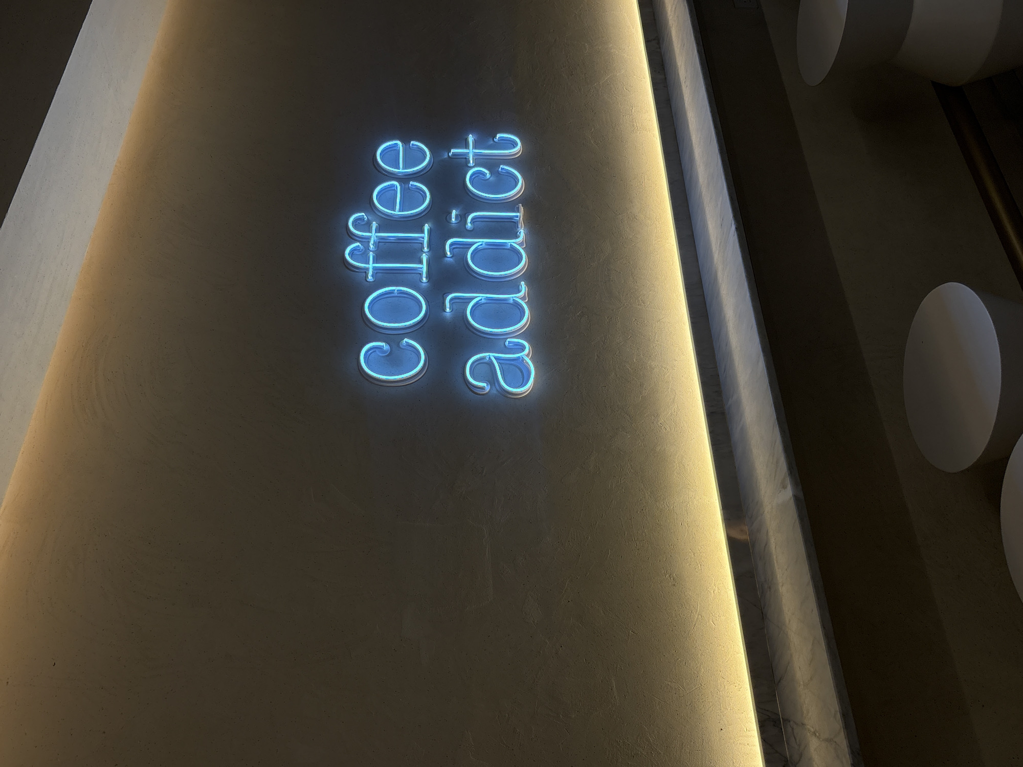 The image shows a wall with the words "coffee addict" written vertically in blue neon lights. The wall is painted in a light beige color. To the right of the wall, there is a narrow vertical strip of light and a row of white cylindrical objects. The location is not explicitly stated in the image and cannot be determined from the available visual information.