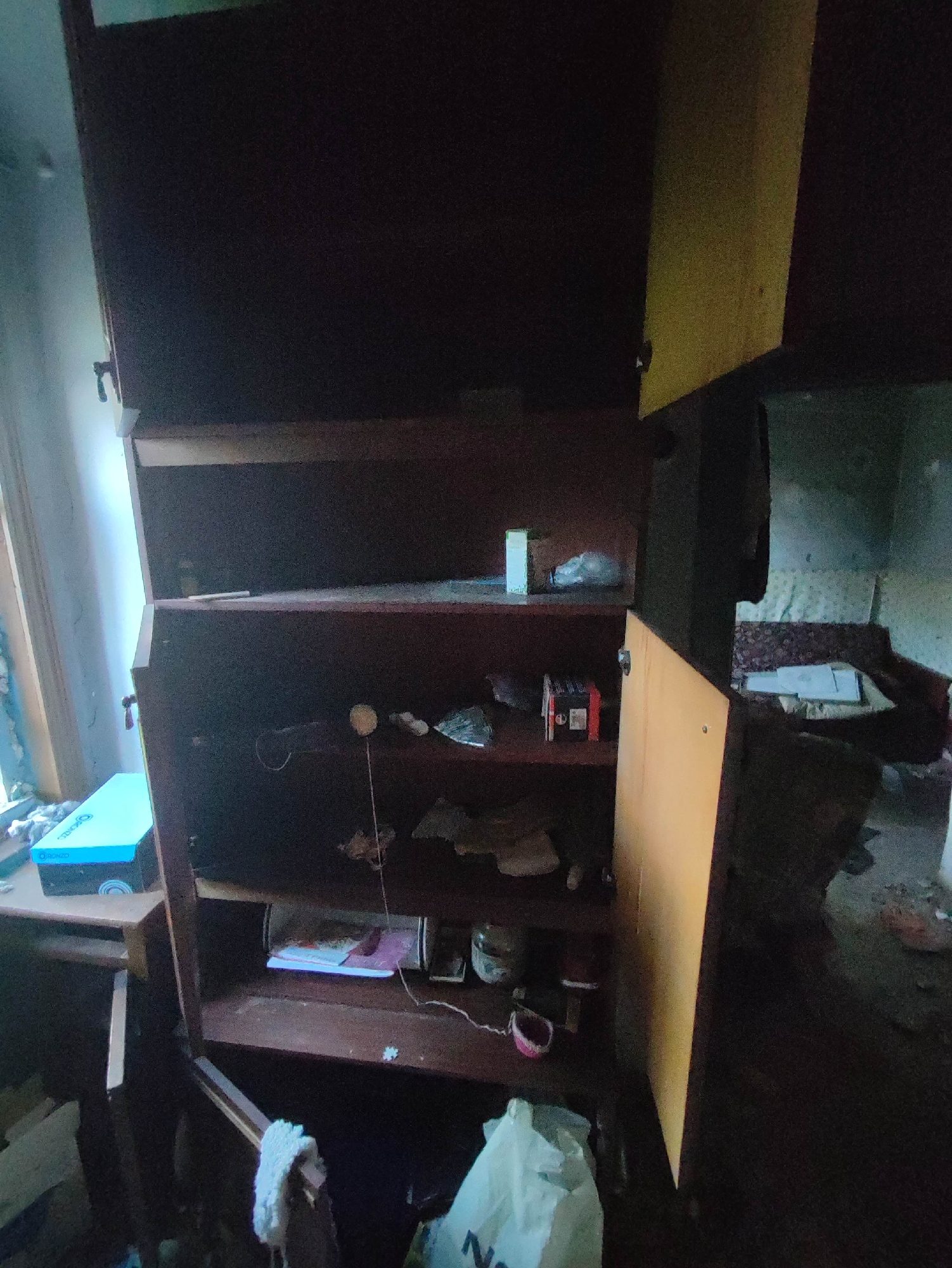 The image depicts a cluttered, dilapidated interior space, possibly an abandoned room in Gremyachinsk, Russia. A large, dark wooden cabinet with open shelves dominates the center of the frame. The shelves are filled with various objects, including what appear to be boxes of ammunition, papers, small containers, and other unidentifiable items. To the left, a blue box with the branding "ORIONZO" is visible on a desk. The room appears generally disorganized and neglected, with visible dust and debris on surfaces and the floor. The lighting is dim, suggesting natural light from a window on the left side of the image. The overall impression is one of disuse and decay.