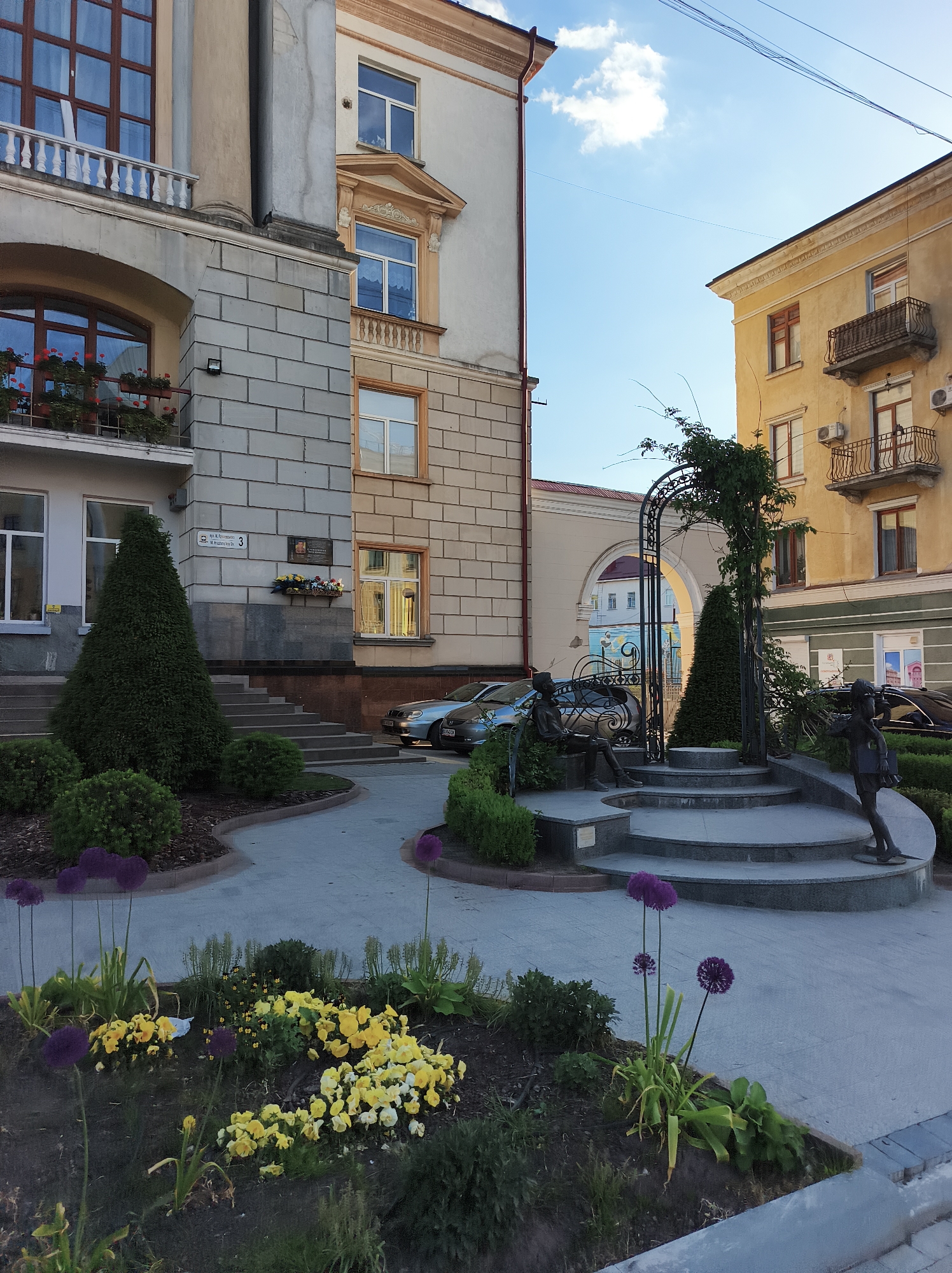 The image depicts a public square in Ternopil, Ukraine. The foreground features a flowerbed with yellow and purple flowers. In the center of the square is a tiered concrete structure with a metal arch covered in climbing plants. Bronze sculptures of two figures are positioned on either side of the structure. In the background, cars are parked in front of buildings with visible architectural details such as windows, balconies, and columns. The building at "3 вул. M.Hrushevskoho" can be seen on the left. The sky is partially visible.