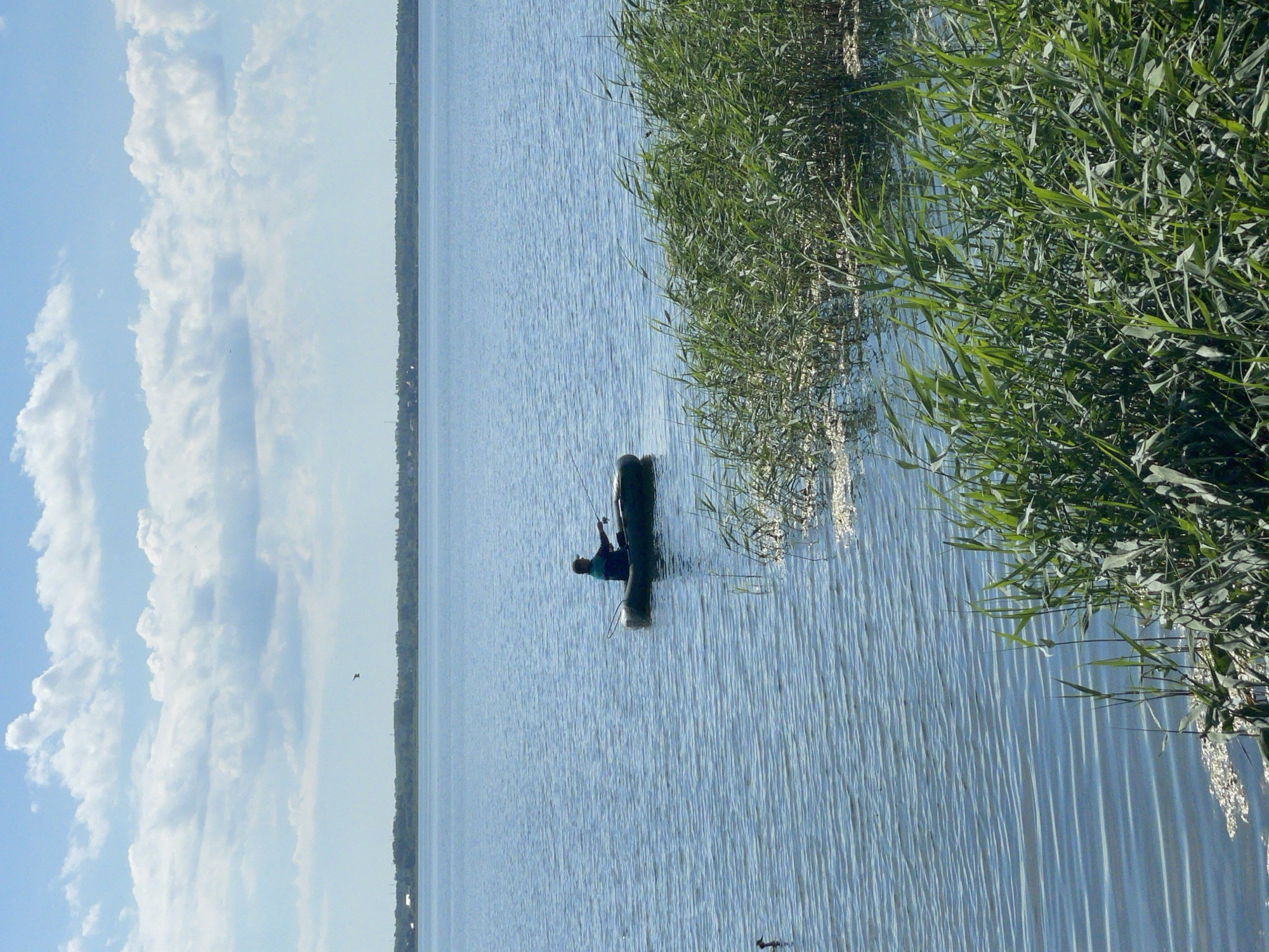 A lone fisherman in an inflatable boat casts a line into the calm blue water of a large lake under a partly cloudy sky. The fisherman is wearing a dark shirt and hat. Dense green reeds line the foreground, and a distant, treed shoreline with scattered buildings can be seen on the horizon. A small bird is visible in the water to the left of the boat.