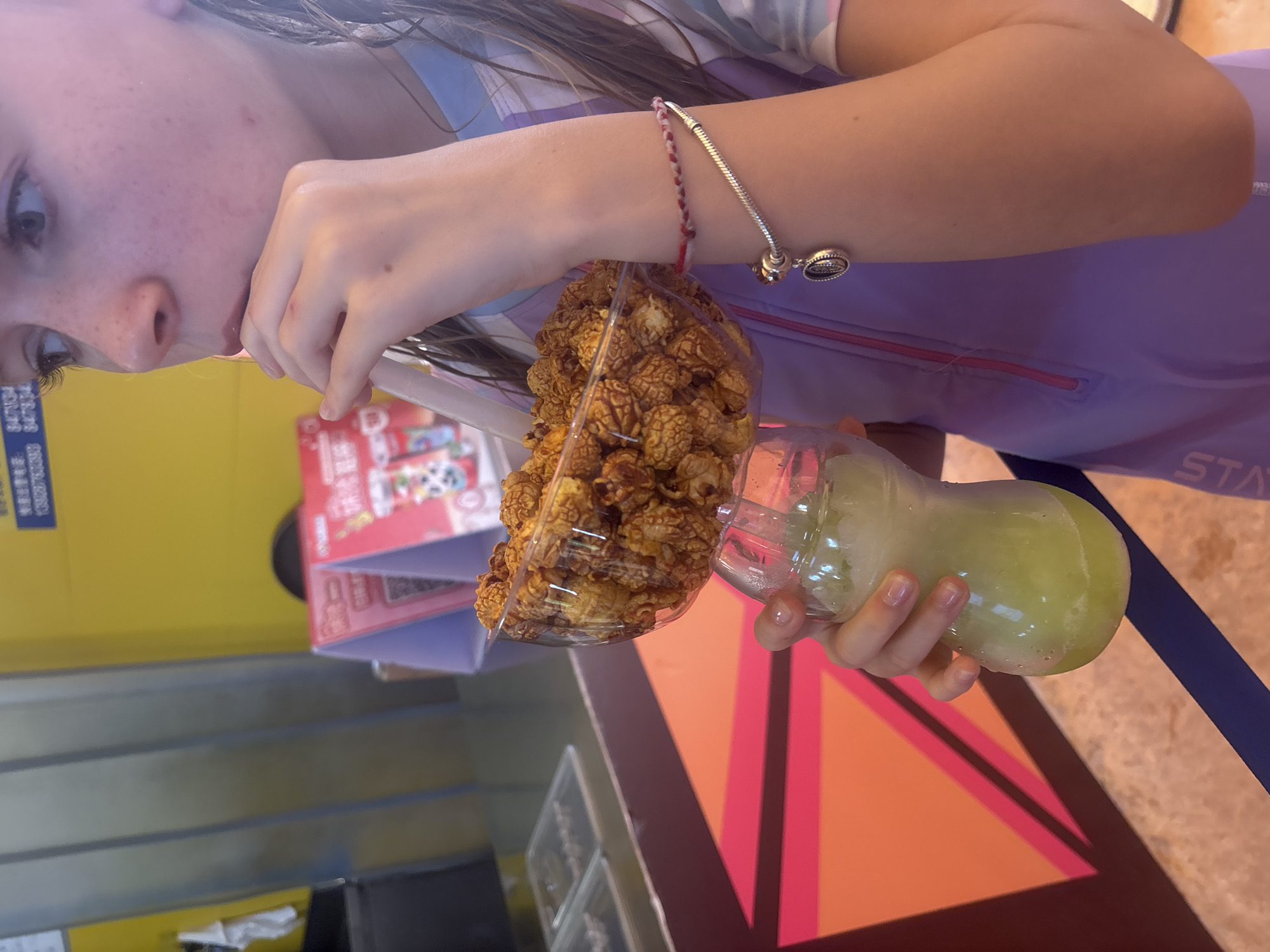 A young person is holding a clear plastic cup filled with caramel popcorn and another clear cup containing a light green beverage. The person's face is partially visible, with freckles and fair skin. They are wearing a purple jacket with a pink zipper. A silver charm bracelet is on their wrist, with two intertwined red and white strings attached to it. The background is a yellow wall with some folded red and white brochures. The scene appears to be indoors, possibly in a commercial establishment. The overall impression is casual and perhaps a moment of snacking.