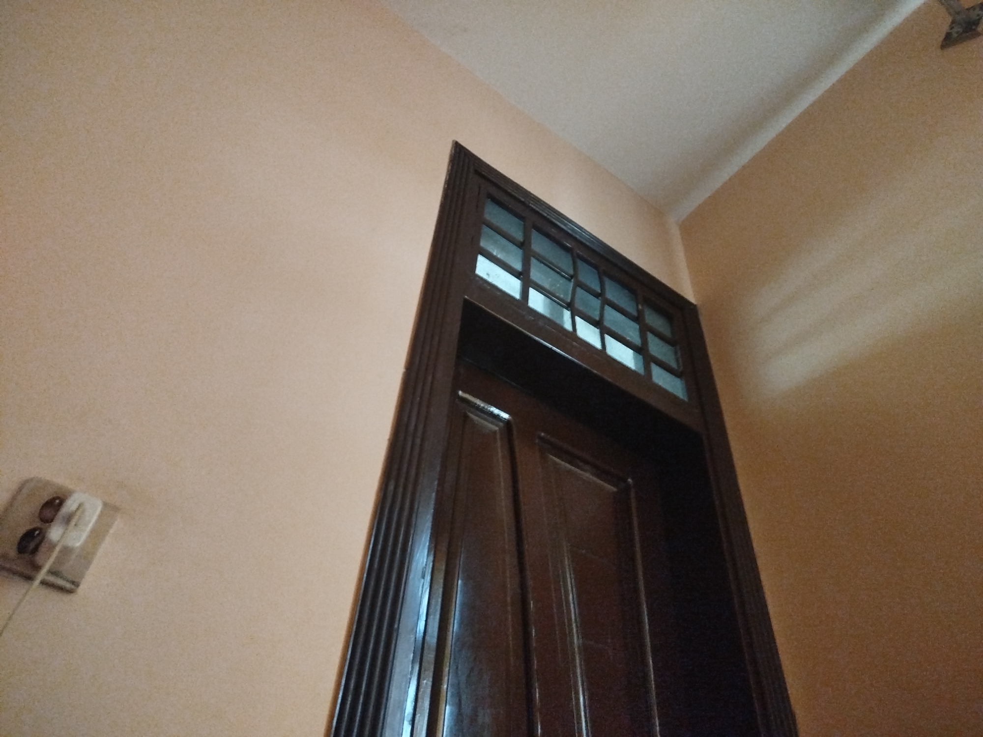 An interior shot shows a dark wooden door with a small window at the top. The door is framed by fluted molding. The wall is painted a light peachy color. To the left of the door, a white electrical outlet with two brown circular objects inside is visible. The ceiling is white and slopes down from the top right of the frame. There is no visible sign of the city of Lassan, Pakistan, or any people.