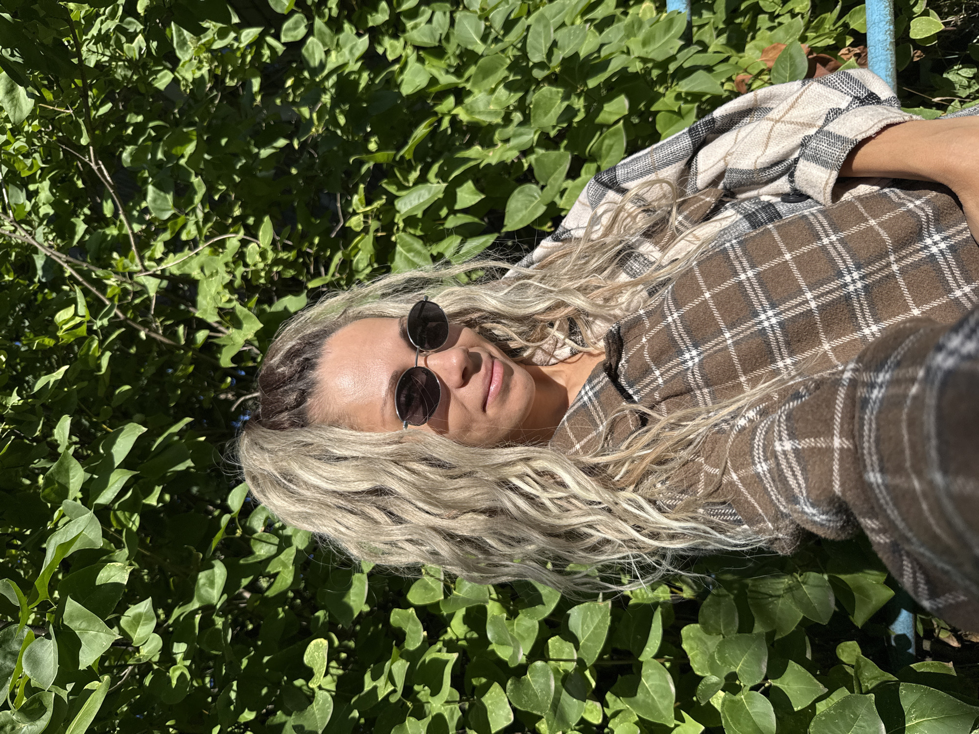 A woman with long, blonde, curly hair tied in a ponytail is pictured from the chest up, facing the camera. She is wearing round, dark sunglasses and a brown and tan plaid shirt. She is standing in front of a dense green leafy bush. The lighting suggests it is daytime with direct sunlight. The image captures a snapshot of the woman outdoors, with the natural environment as the backdrop. The specific location in Rusanovo, Russia is not discernible from the visual information provided in the image.
