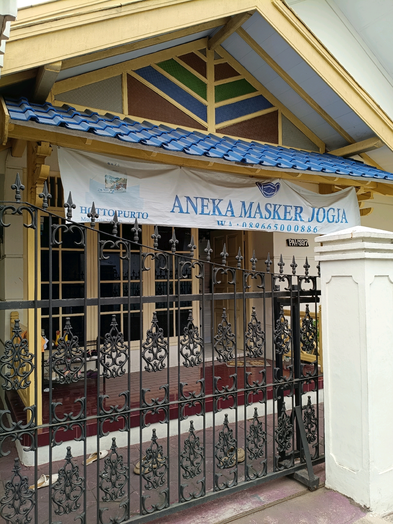 The image shows the exterior of a building in Yogyakarta, Indonesia, with a white banner displaying "ANEKA MASKER JOGJA" and a phone number. The building has a yellow-painted facade and a blue tiled roof. A black wrought-iron gate with ornate designs stands in front of the entrance. Some sandals are visible on the ground behind the gate. The architectural style suggests a residential or small commercial building typical of the region.