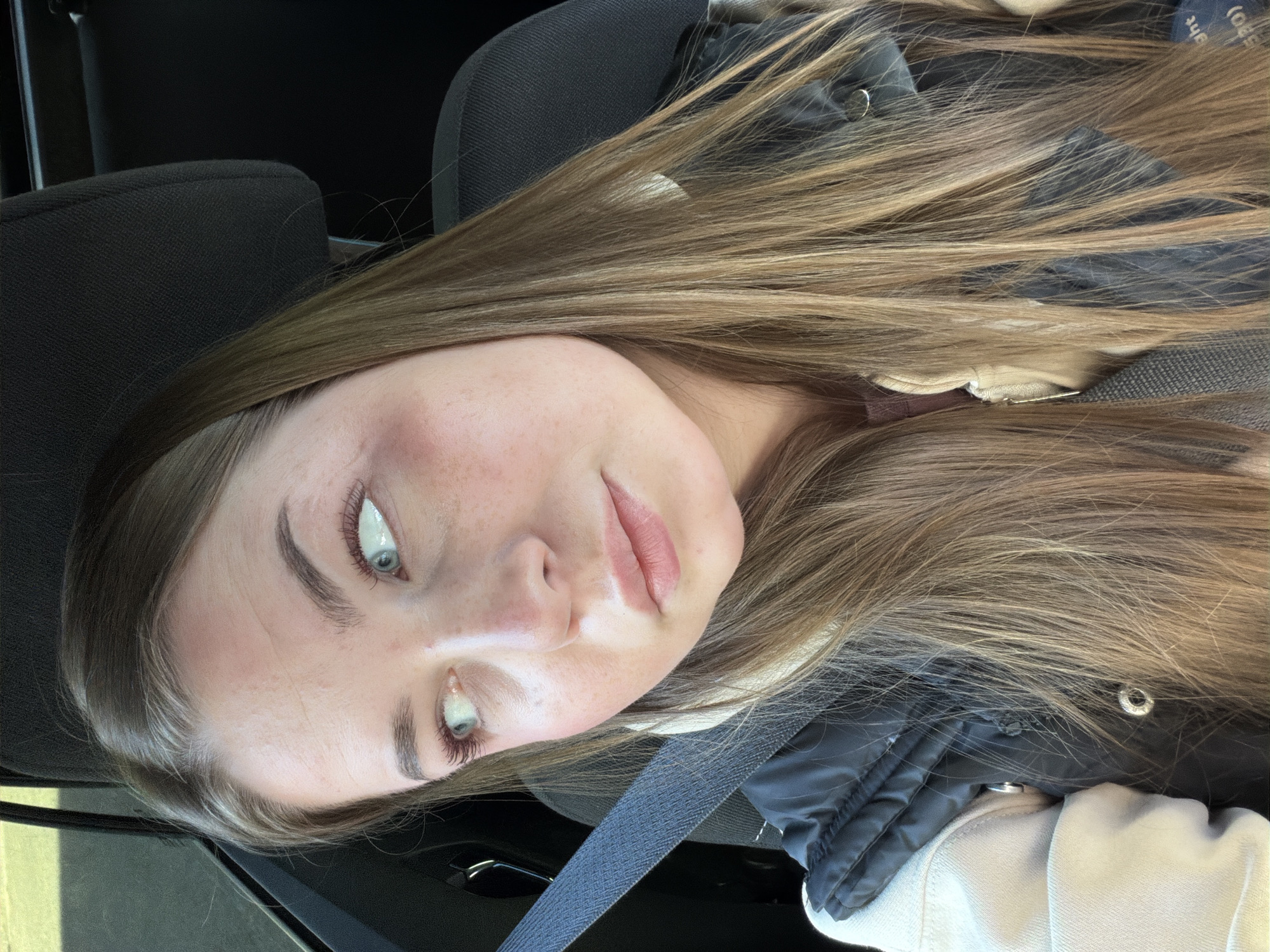 A fair-skinned young woman with long brown hair sits in a car wearing a black and white jacket and a seatbelt. She is likely the driver or front passenger based on the positioning of the seatbelt. The car's interior appears dark. There are no other people, actions, or interactions visible. The location, Chaikovsky, Russia, cannot be determined based on the visual information provided.