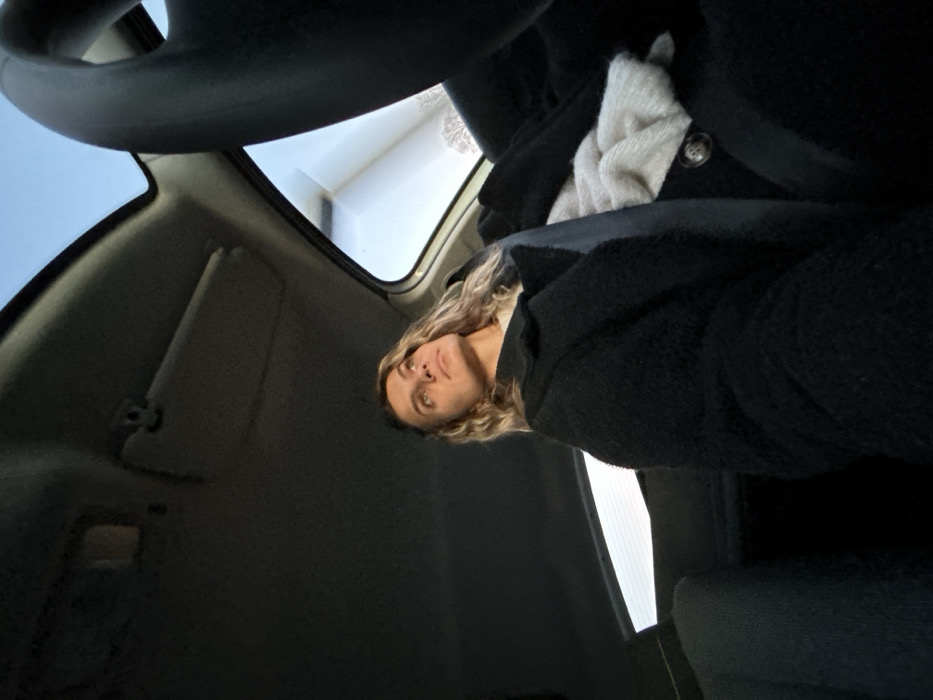 A woman with blonde wavy hair is captured from a low angle inside a car. The steering wheel dominates the upper part of the frame. The woman is wearing a dark, textured coat and a light-colored scarf. The car's interior features a sun visor, windshield, and side window. Light enters through the windshield, creating a contrast within the car's cabin.