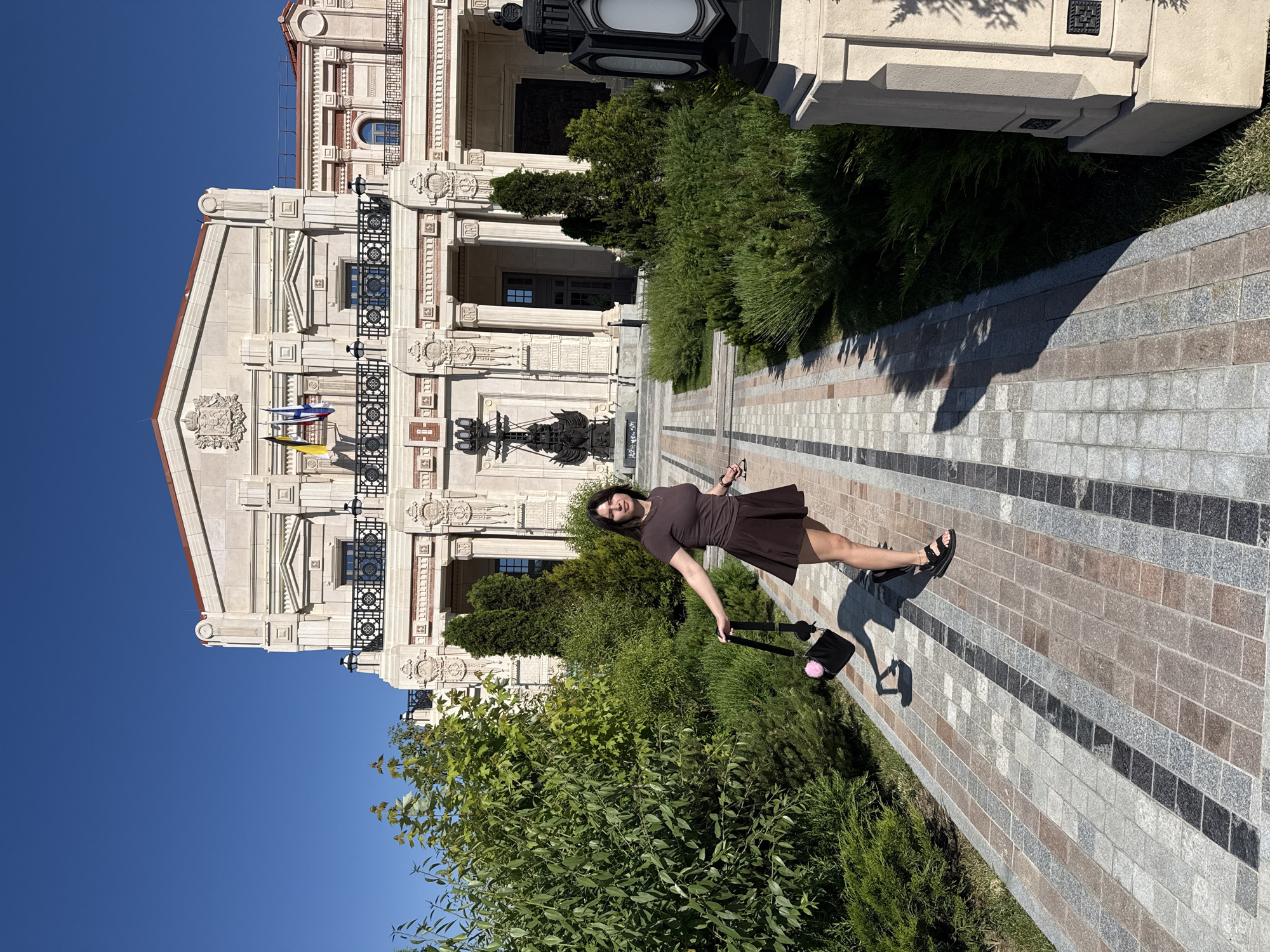 In the city of Unknown, Unknown, a young woman stands on a paved walkway in front of an ornate building. She is wearing a brown short-sleeved dress and a matching short skirt, along with black sandals. In her left hand, she holds a black handbag with a pink pom-pom. The building behind her is grand, featuring classical architectural elements like columns, balustrades, and decorative carvings. Flags are visible on the upper balcony. Lush green bushes and trees surround the walkway, creating a garden-like setting. The sky is clear and blue. The image is taken during the daytime, with the sun casting shadows on the ground.