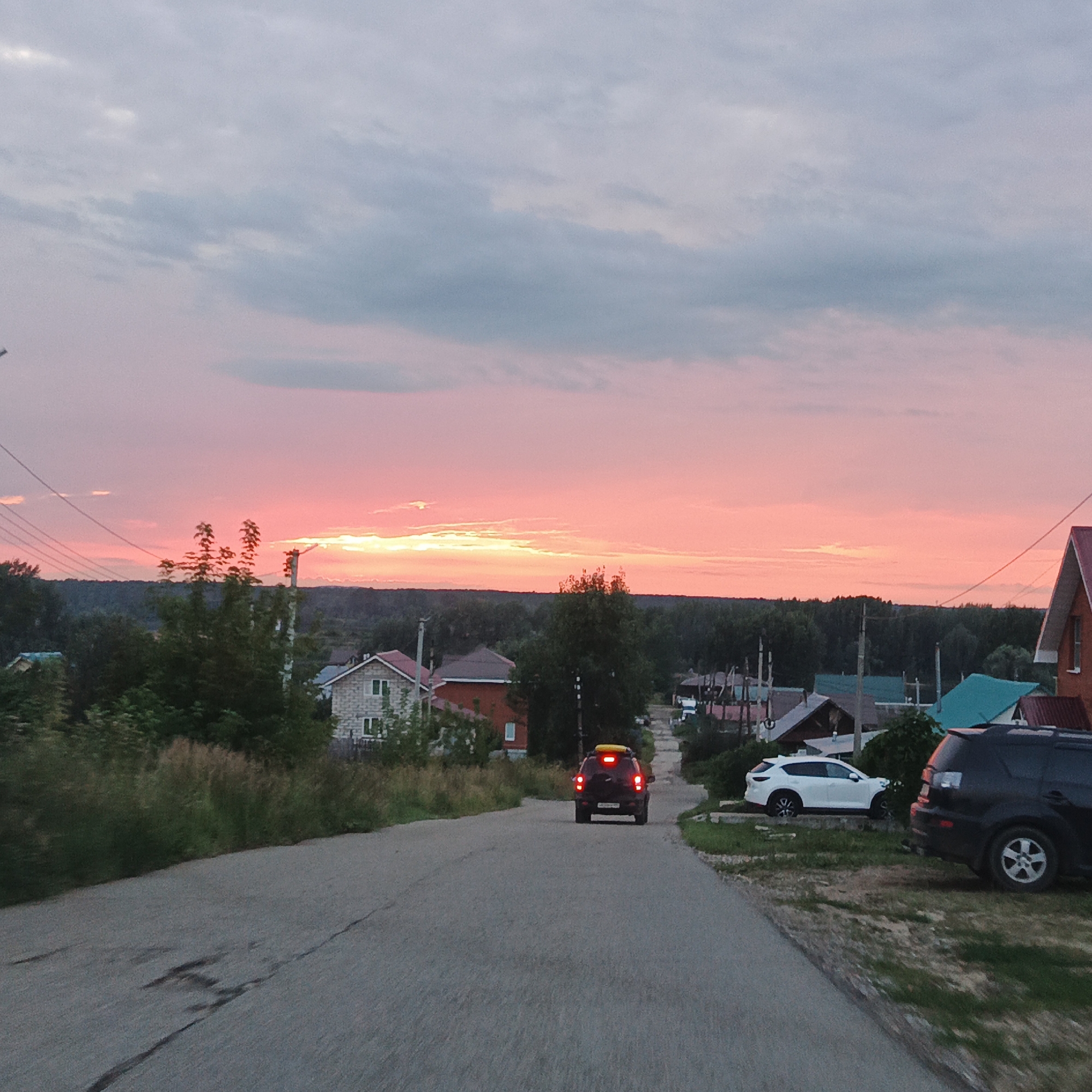 An asphalt road curves through a rural village during a sunset with a vibrant pink and orange sky. In the foreground, the road is cracked and gray. A dark SUV with its brake lights illuminated is driving away from the viewer up the road. To the right of the road, a white SUV is parked. In the middle ground, several houses with pitched roofs are visible amidst trees and tall grass. In the background, a dense line of dark green trees forms a horizon against the colorful sky, which is filled with soft, gray clouds. Overhead power lines stretch across the sky.