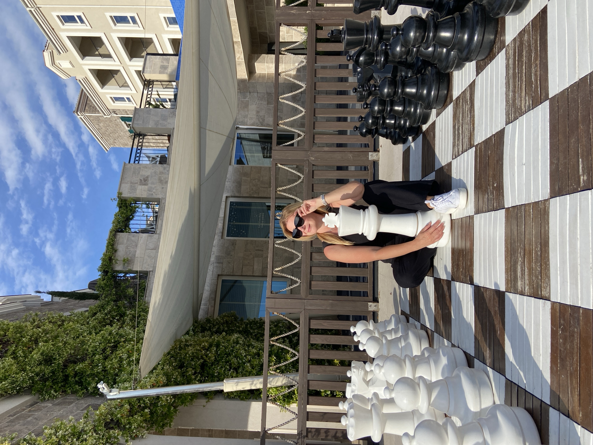 A woman with blonde hair, wearing sunglasses, a black top, and black pants, is posing on a large outdoor chessboard. She is holding an oversized white queen chess piece with both hands, looking towards the camera. Her white-sneaker-clad left foot rests on a white square of the board.
The chessboard itself is constructed from alternating dark brown and white wooden planks. On one side, a full set of large black chess pieces is arranged, while several white pieces, including pawns, rooks, knights, bishops, and a king, are visible on the other side. These pieces cast long, distinct shadows, indicating bright sunlight.
In the background, multi-story resort-style buildings with balconies are visible under a clear blue sky dotted with scattered white clouds. The buildings feature light-colored facades and dark metal balcony railings. Green climbing plants and bushes cover portions of the buildings and extend along wooden fences, which incorporate rope detailing. Reflections, possibly showing "Coffee Bay" and operating hours like "08.00-18.00," are faintly visible in a window, suggesting a cafe or similar establishment within the complex.
The scene takes place outdoors on a sunny day, likely midday or early afternoon given the sharp shadows, in what appears to be a recreational area of a resort or hotel. The location context provided is Radovići, Montenegro.