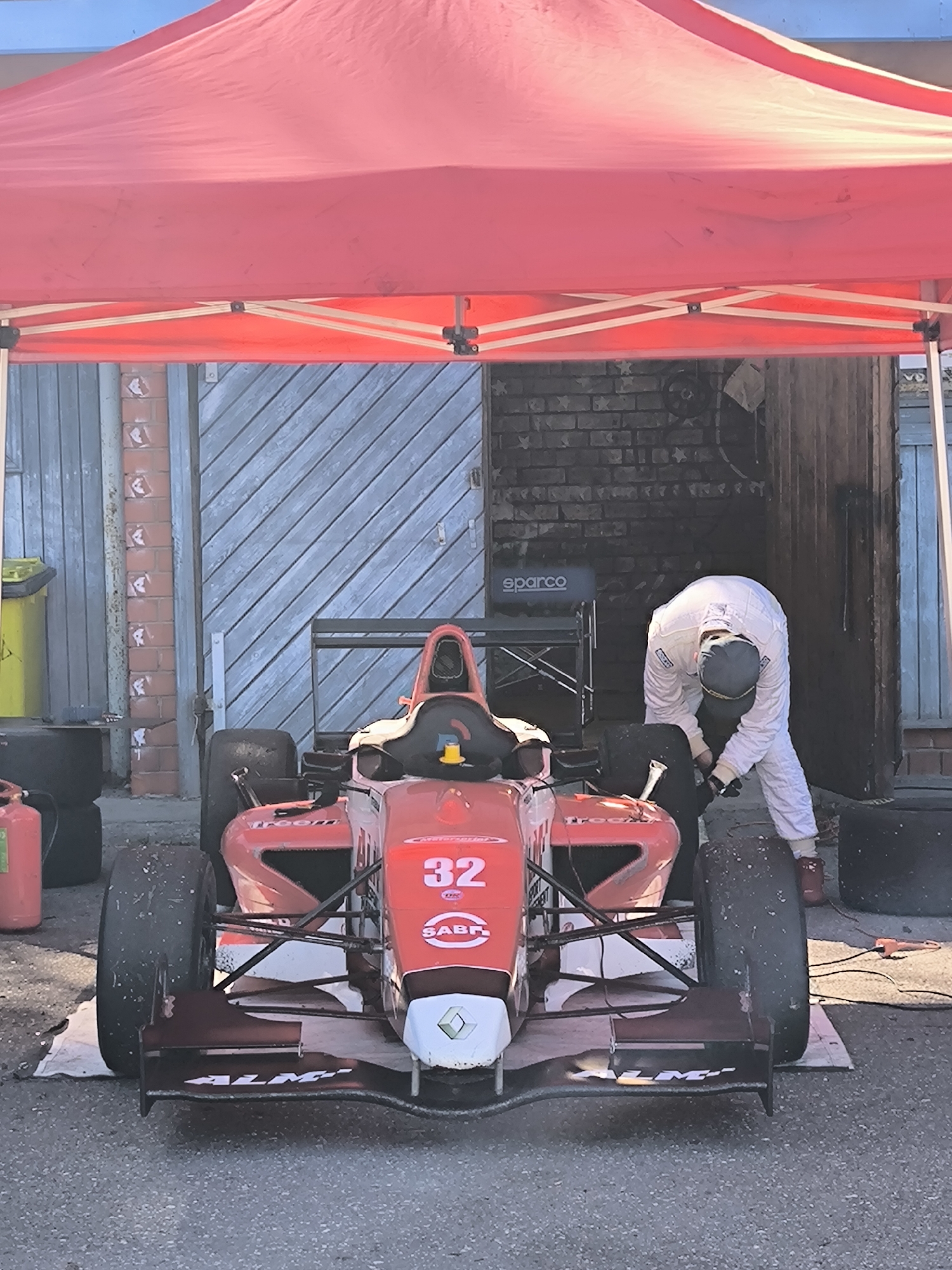 The image depicts a red Formula race car, numbered "32," positioned under a red canopy. A person wearing a white jumpsuit and a dark cap is attending to the car, appearing to work on the tire. The background includes a building with a gray, diagonally-paneled door and a brick section with what appear to be decorative squares. The presence of "sparco" branding on a chair behind the vehicle and safety equipment like a fire extinguisher indicates a racing support environment. The location, Riga, Latvia, is not evident in the image without external information.