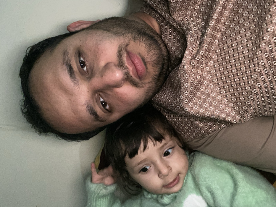 An upward-facing, close-up portrait shows a man and a young child, likely a father and daughter, lying down together. The image is taken from above, with both subjects looking towards the camera, suggesting a selfie. The man, positioned in the upper part of the frame, appears to be an adult with dark hair and a short beard. He is wearing a brown patterned shirt, possibly a traditional kameez. His expression is calm and neutral, with a slight hint of a smile.
Below and slightly to the right of the man is a young child, appearing to be a toddler, with dark hair and eyes. She is wearing a light green, possibly fuzzy or knitted, long-sleeved sweater. Her gaze is directed slightly off-camera to her right, and she has a subtle, curious expression.
The background is a plain, light-colored surface, possibly a wall or bedding, indicating an indoor setting. The image lacks distinct location cues specific to Kandahar, Afghanistan, other than the general appearance of the individuals and the man's attire which could align with regional clothing styles. There is no visible text. The overall mood is intimate and peaceful.