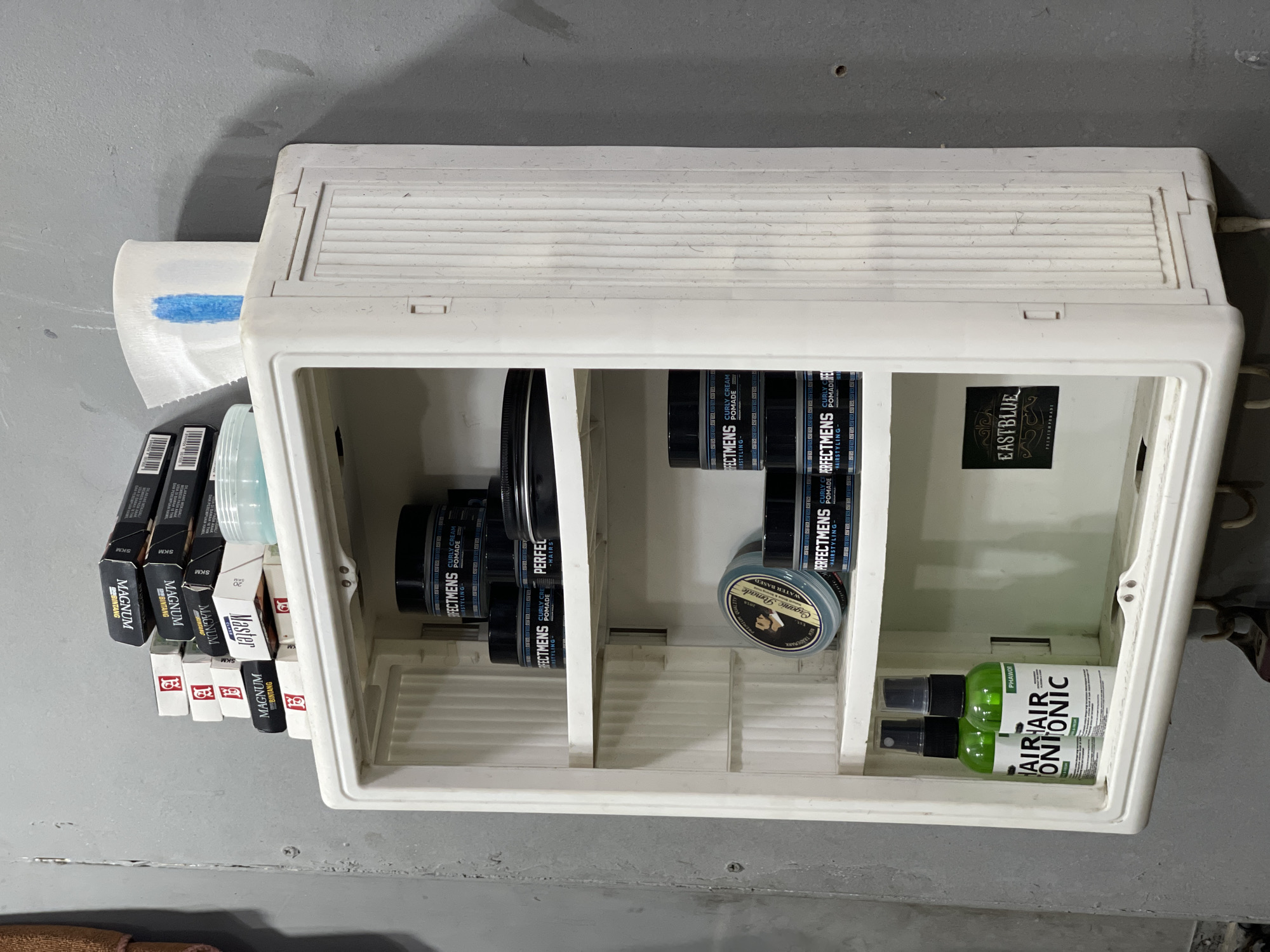 An indoor, eye-level, medium shot captures a white plastic cabinet mounted on a gray concrete wall. The cabinet is open, revealing three shelves. The top shelf holds a stack of cigarette packs, including "Magnum Bintang" and "Master," alongside a partially visible blue and white roll of paper towel and a small blue jar. The second shelf contains several black jars labeled "Perfectmens Curly Cream Pomade." The third shelf displays a round tin of "Organic Pomade" and more "Perfectmens" jars. The bottom of the cabinet holds two green spray bottles labeled "Hair Tonic." A black sticker with "EASTBLUE PREMIUM POMADE" is visible on the back wall of the cabinet. The overall scene is static and lacks human presence. The environment appears to be a storage or grooming area.