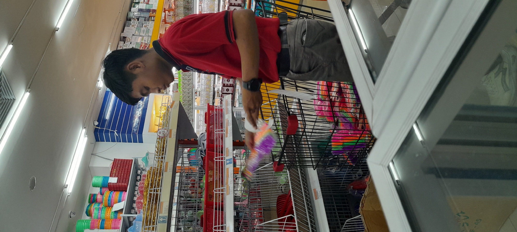 A young man wearing a red polo shirt and grey pants is standing in a brightly lit supermarket aisle, focused on placing a multi-colored plastic item into a shopping cart filled with similar items. The shopping cart is black with red accents. Shelves in the background are stocked with various household goods, including stacked colorful bowls and red plastic baskets. A white cooler with reflective glass is in the foreground. The scene appears to be in a supermarket in Rogojampi, Indonesia.