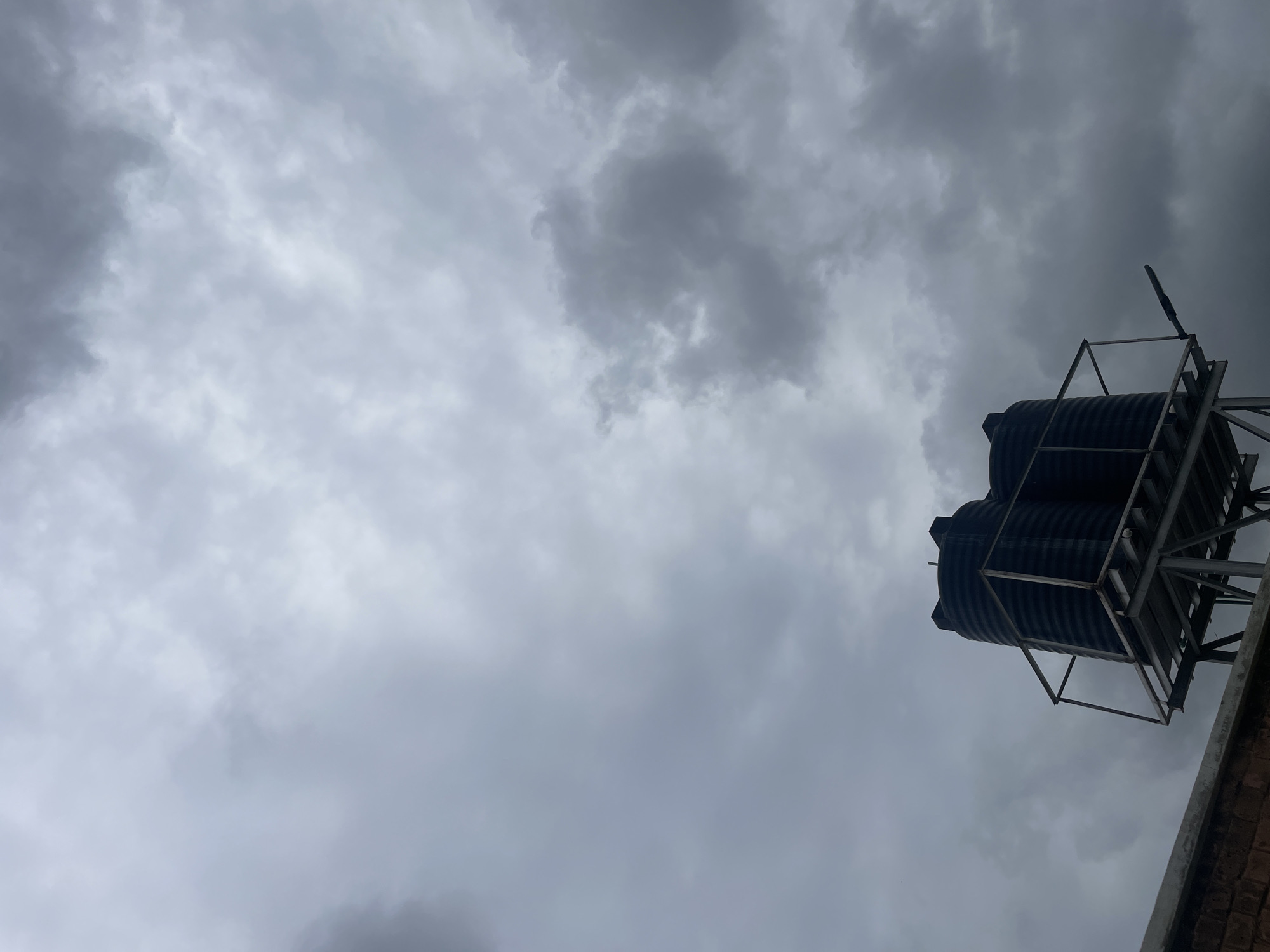 The image depicts a dark, overcast sky filled with heavy clouds. In the foreground, two black water tanks are mounted on a metal stand, situated on the edge of a concrete structure, likely a rooftop. A brick wall is partially visible at the bottom left of the frame. The scene appears to be from Barako, Nigeria, given the context provided. There are no people or animals visible in the image. The overall atmosphere is somber, possibly indicating impending rain.